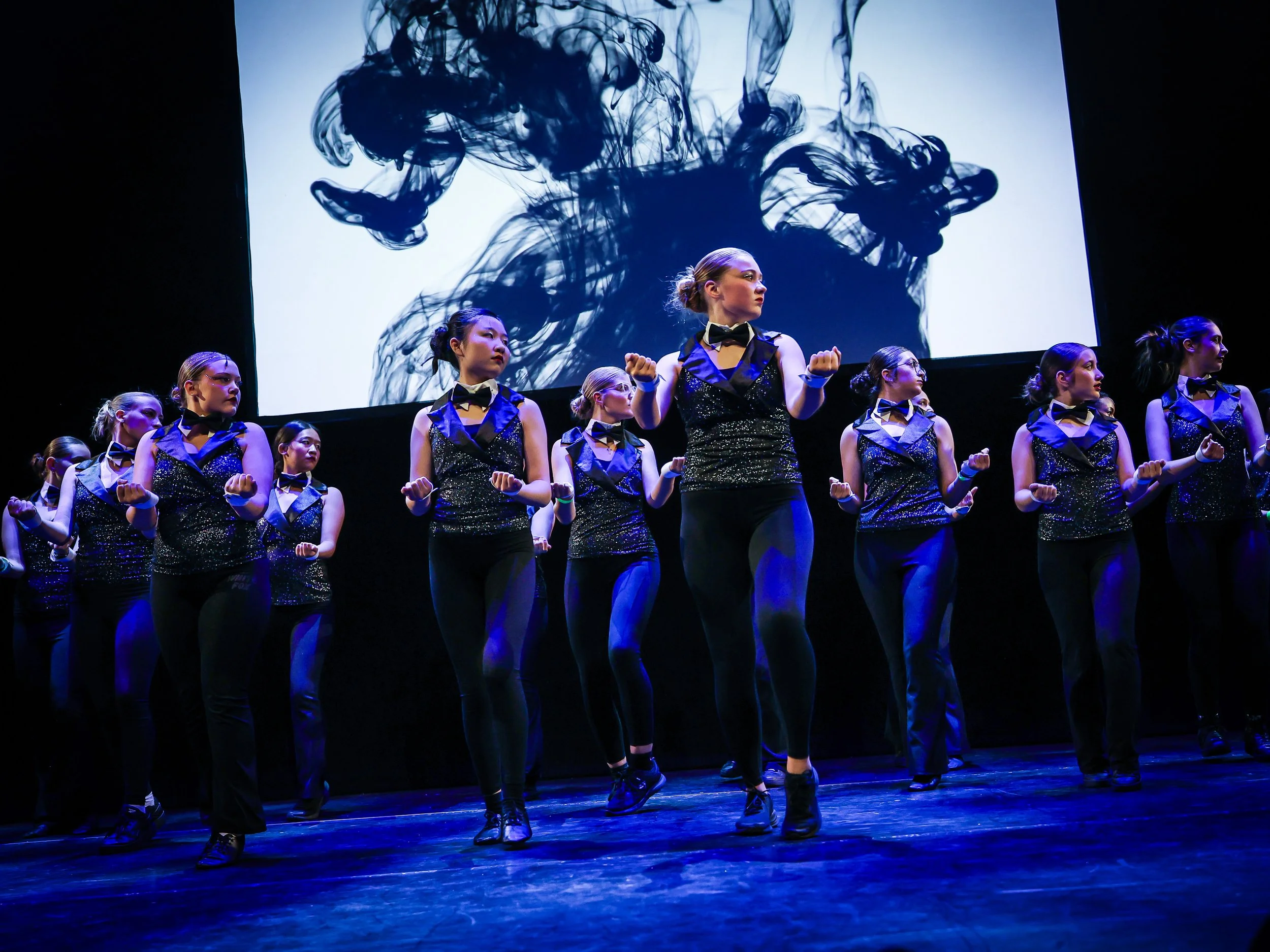 A group of female dancers performing on stage in matching costumes with black tops, blue accents, and black pants, with a black and white abstract visual projected behind them.