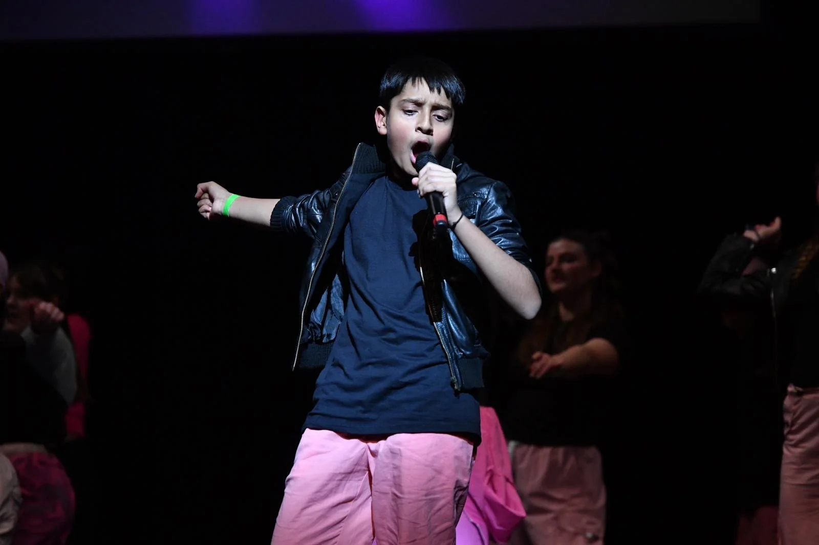 Young boy singing into a microphone on stage, wearing a black leather jacket and pink pants, with other children in the background.