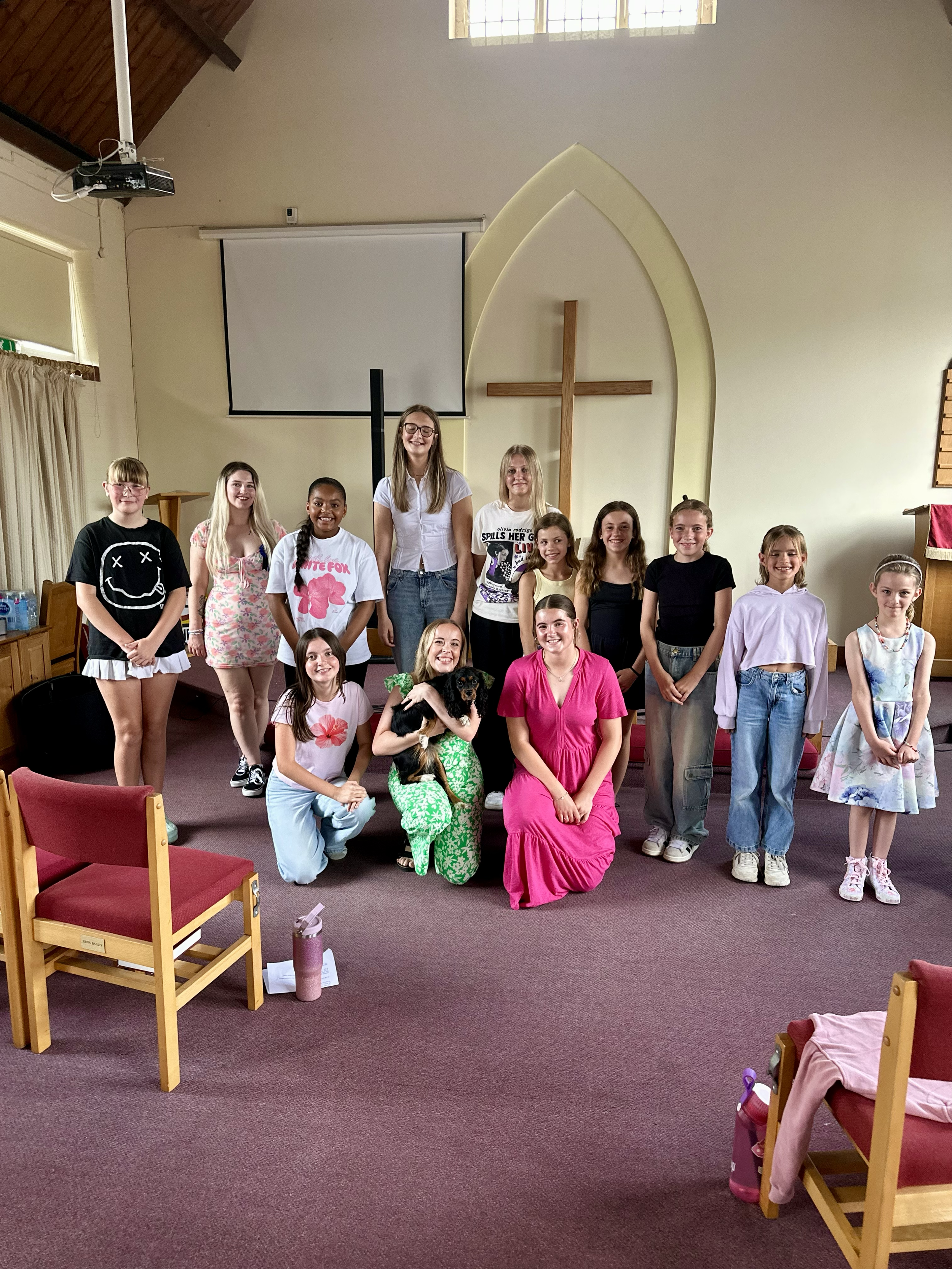 A group of young girls and women, including a woman in a pink dress, posing with a small dog in a church interior with a cross and a large projection screen in the background.
