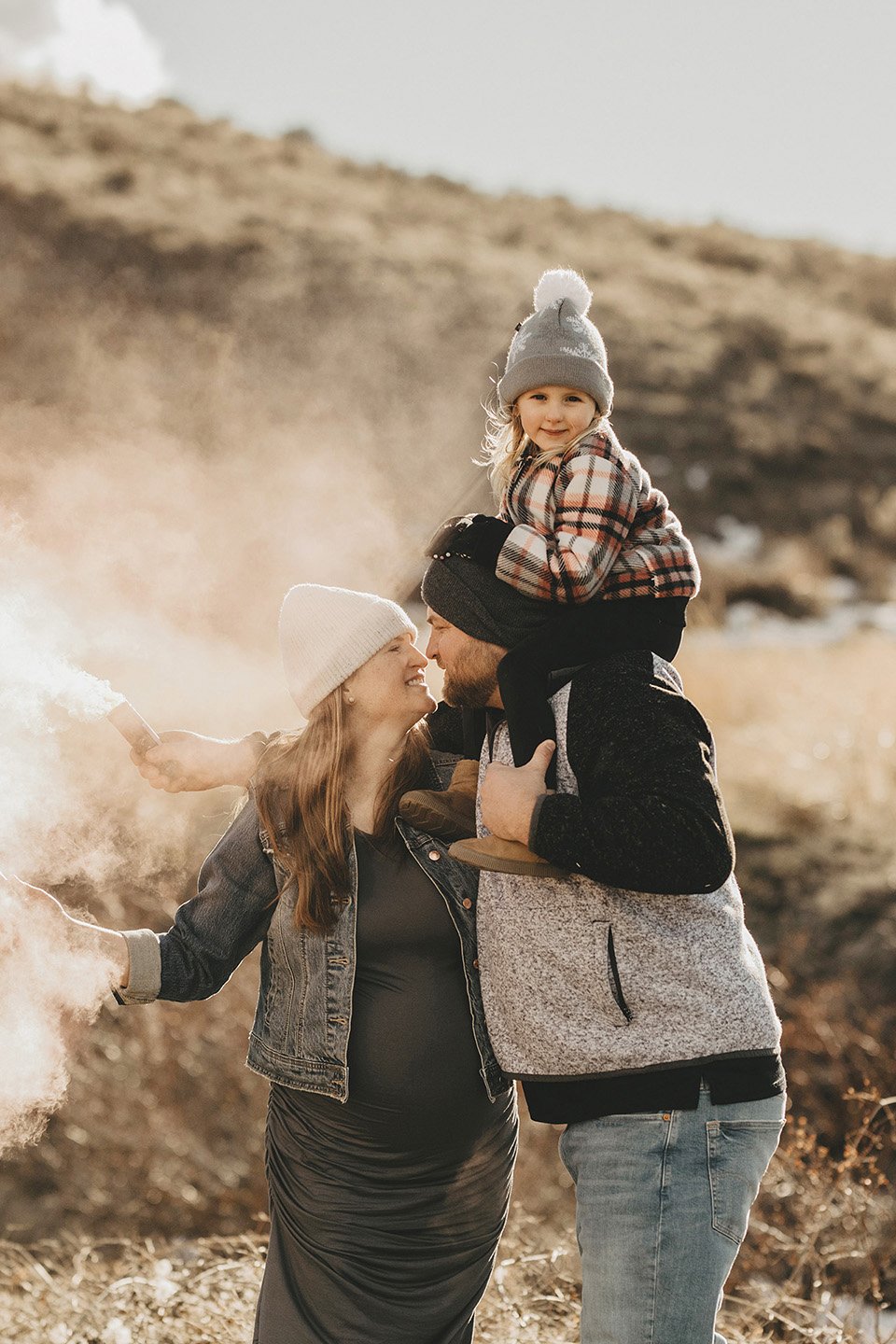 A joyful gender-reveal maternity session in Yakima, where the couple shares a tender nose-to-nose moment as pink smoke fills the air and their daughter beams from dad’s shoulders.