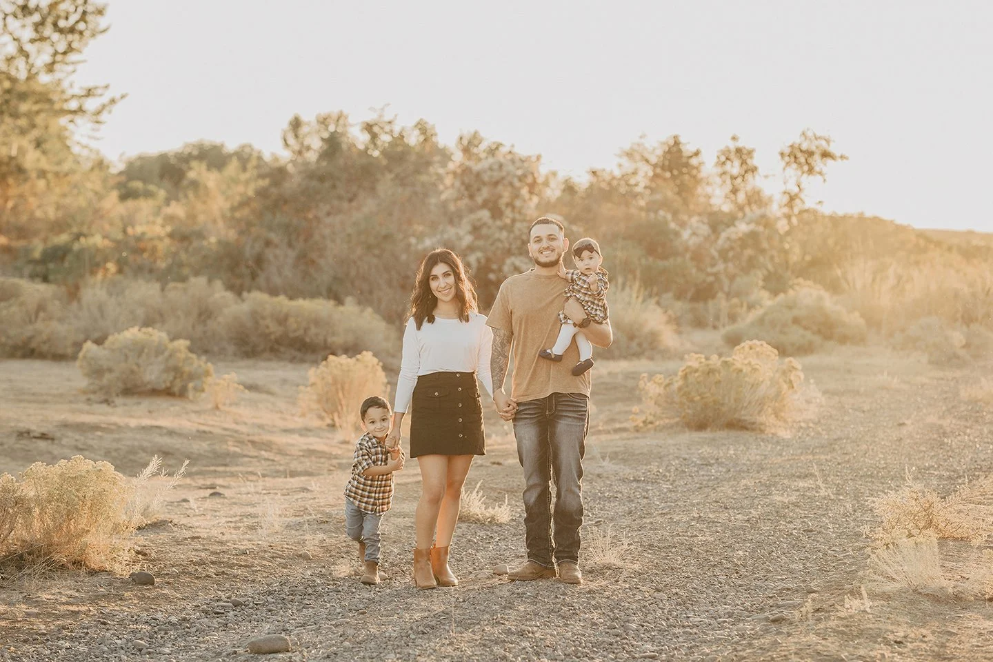 Family of four in Toppenish, WA during golden hour — natural smiles, dad holding the baby, and a sweet moment with the older boy standing close to his mom in a dry, warm-toned location.