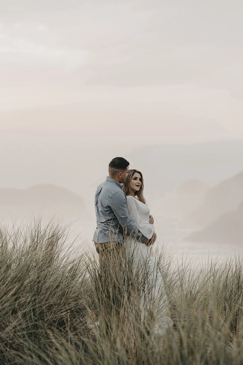 A serene coastal maternity moment at Cannon Beach, where the husband stands lovingly behind his wife as they’re surrounded by tall beach grass, ocean fog, and soft natural tones.