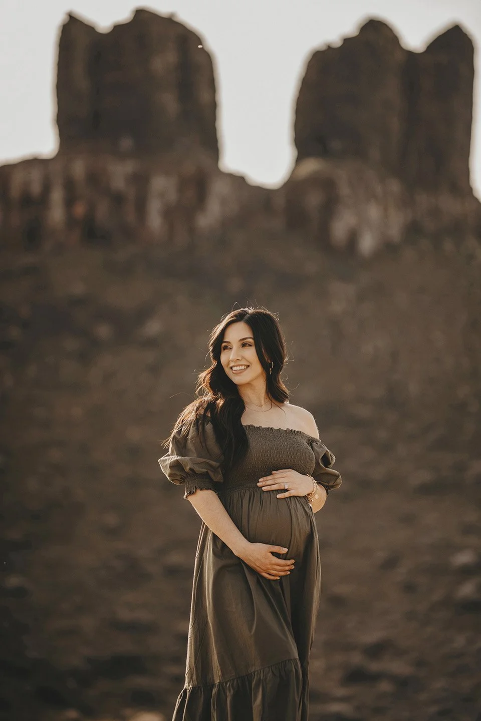 A soft, intimate close-up at Twin Sisters Rocks, where the mother-to-be gently holds her belly in a flowing green dress, surrounded by the calm beauty of spring.