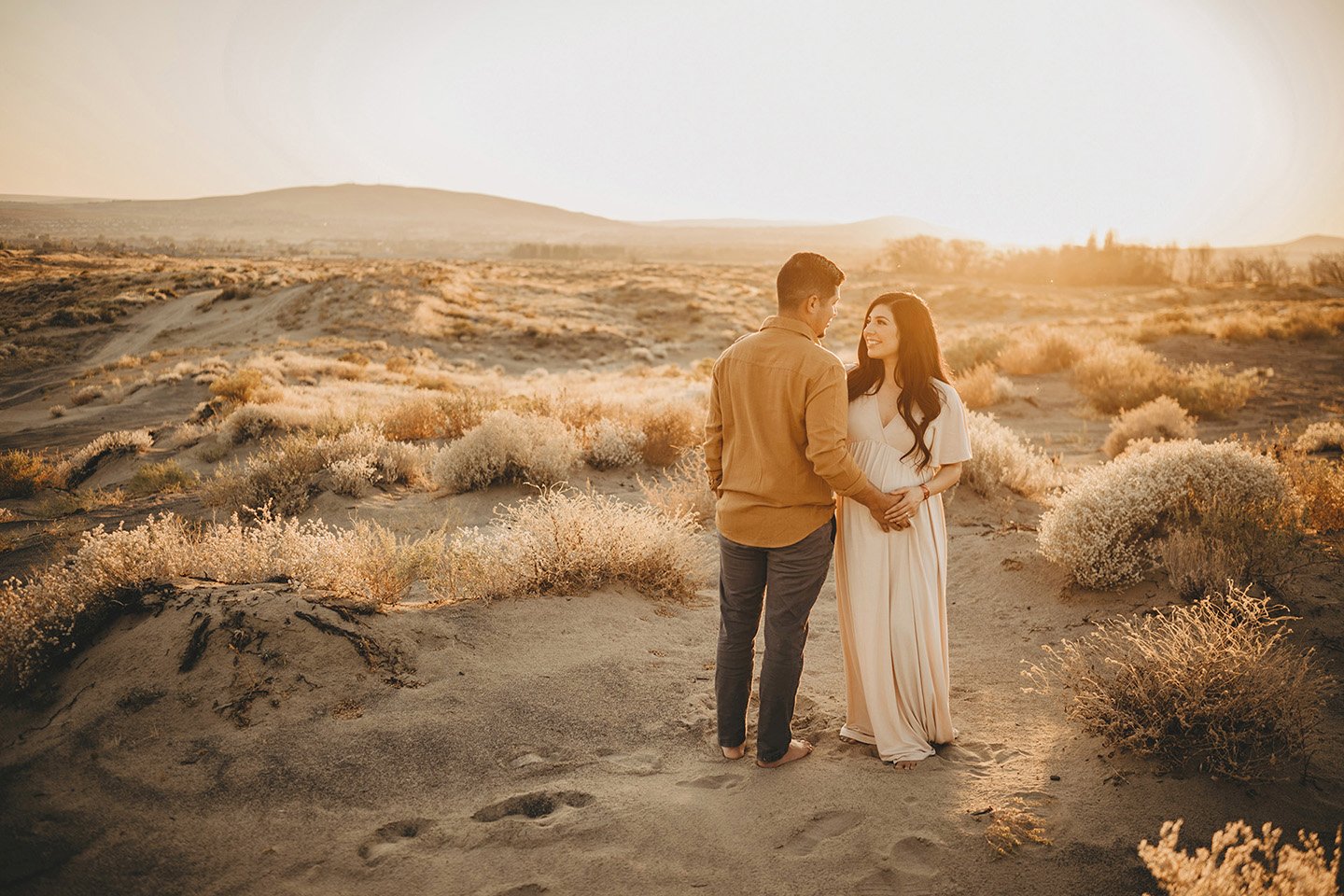 A beautiful full-body maternity portrait at the Richland sand dunes, where glowing golden light frames the couple as they smile warmly at each other.
