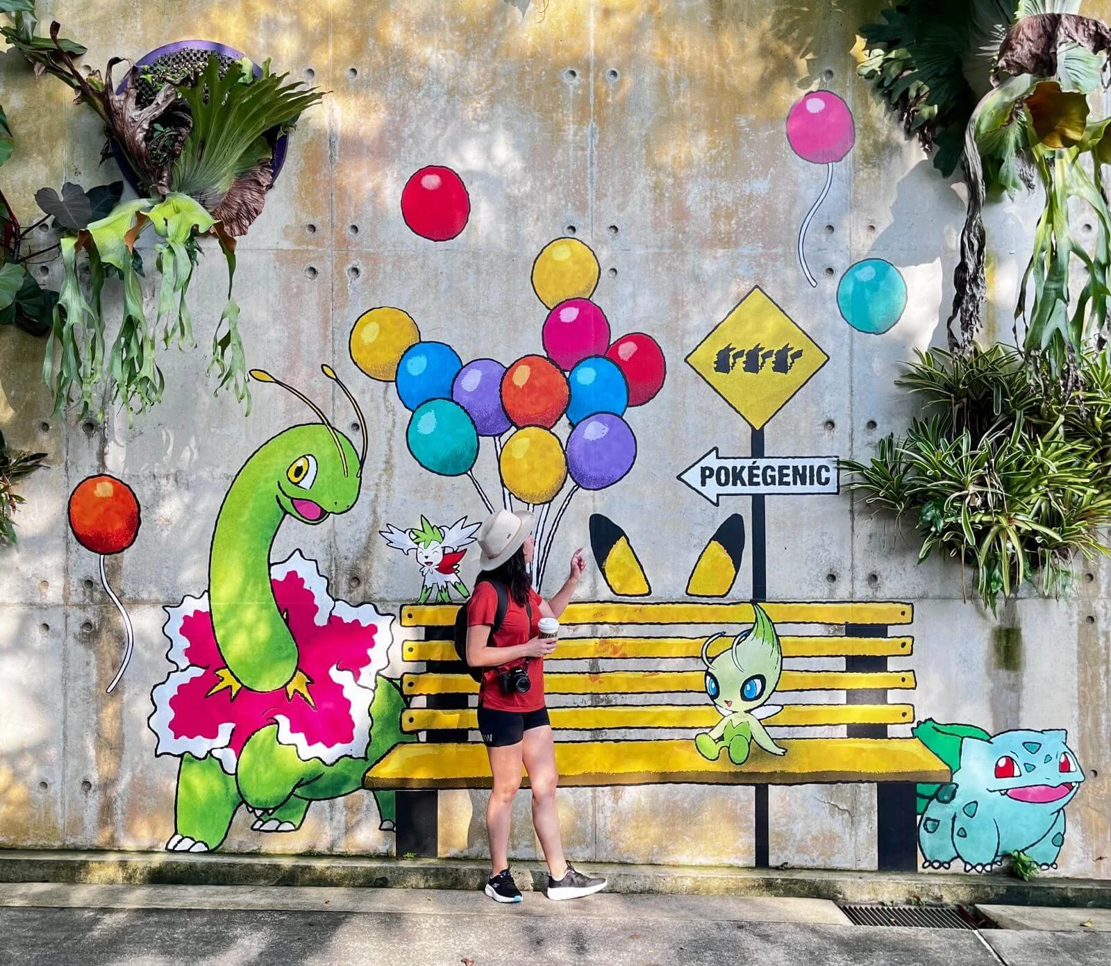 Mural colorido con personajes de Pokémon en una pared de cemento, con plantas en los lados y una persona con mochila y sombrero, sosteniendo una taza, caminando frente a él.