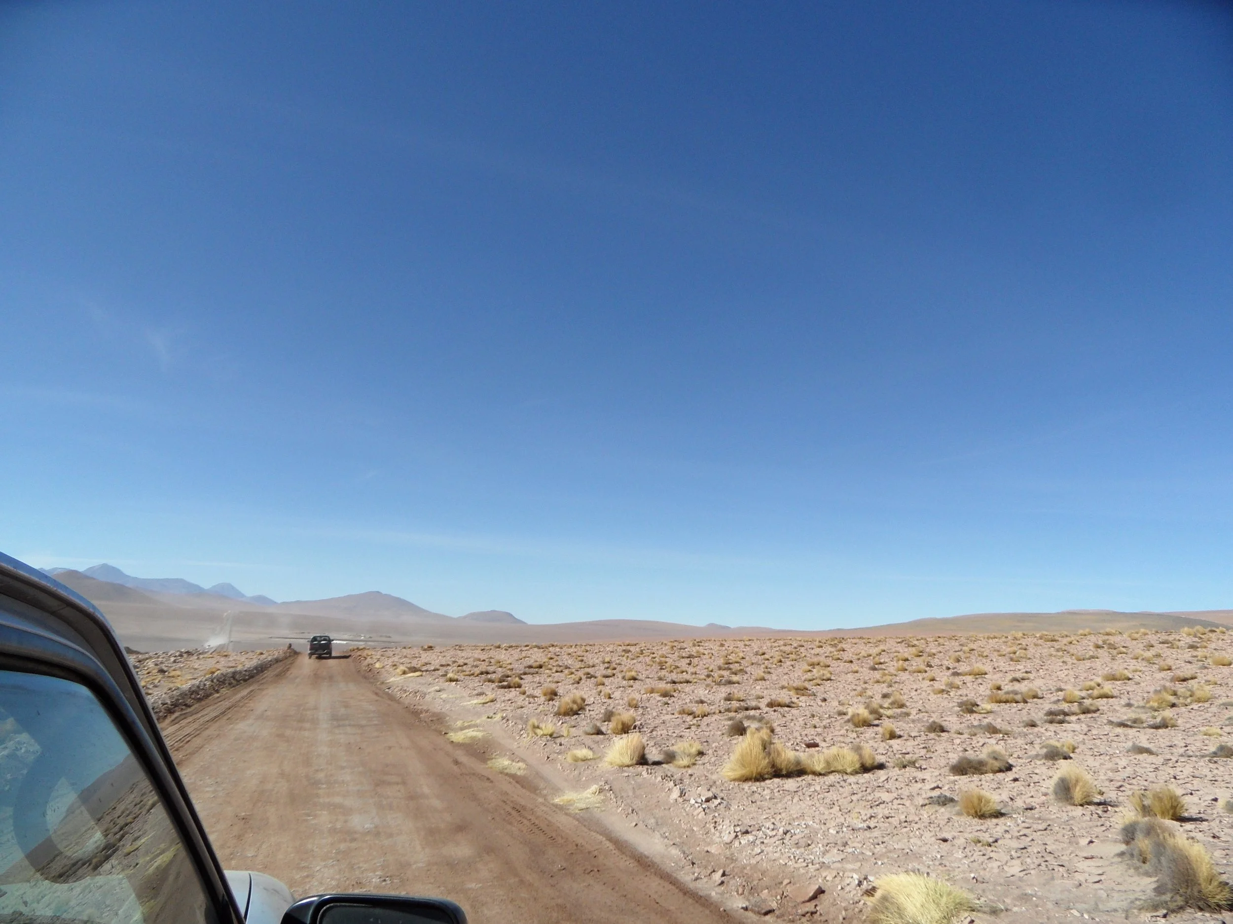 Camino de tierra en un paisaje árido con arbustos dispersos, montañas en la distancia y un cielo despejado.