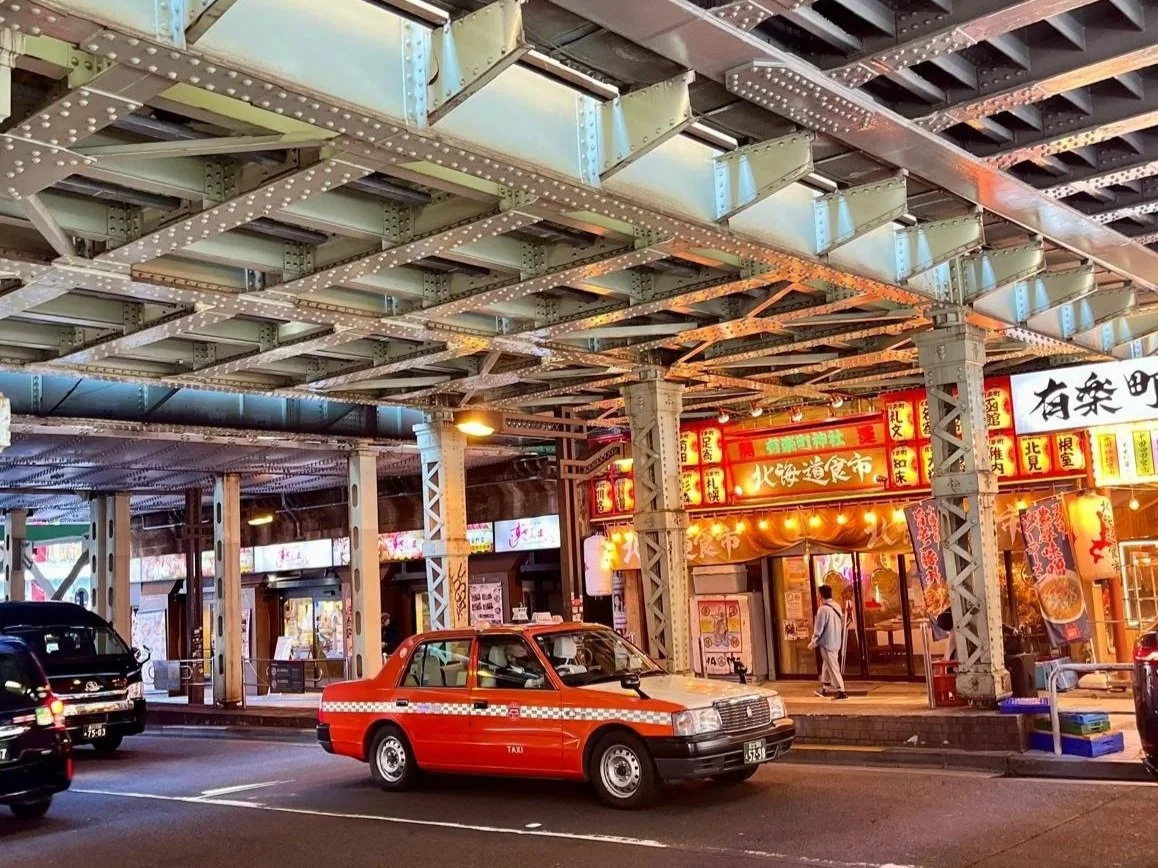 Vista nocturna de un callejón en Japón con un taxi rojo estacionado y tiendas con signos en japonés iluminados.