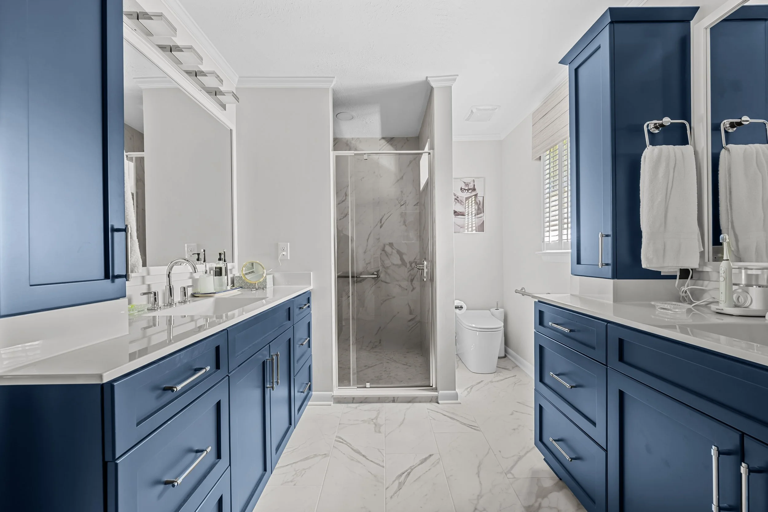 Primary bathroom with opposing blue vanities, tall storage cabinets, marble-look flooring, and a glass-enclosed shower.