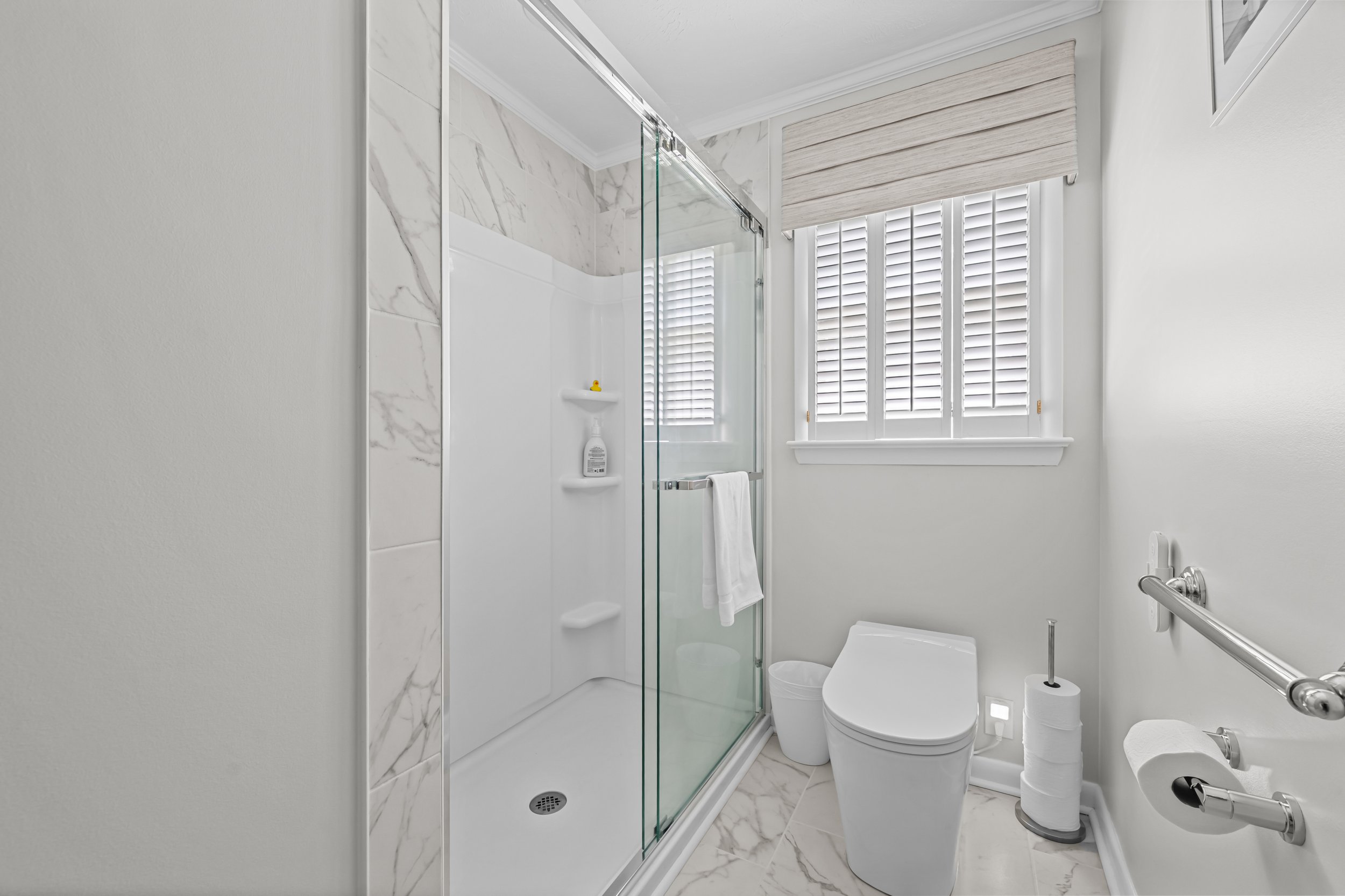 Updated bathroom with a glass-enclosed shower, marble-look wall and floor tile, and grab bars.