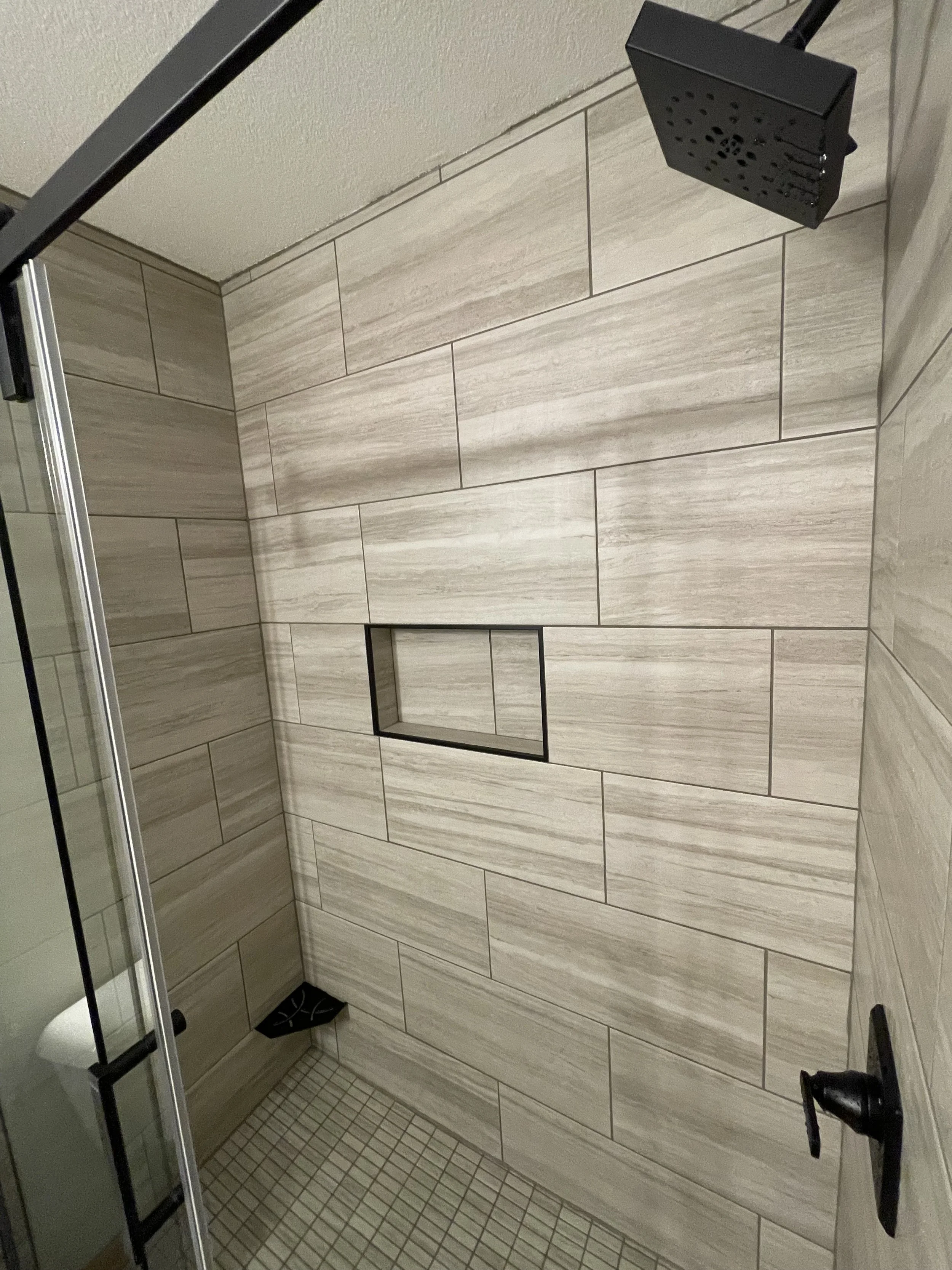 Interior view of a modern shower with beige rectangular wall tiles, a recessed shelf with black frame, a black overhead showerhead, and a glass door.