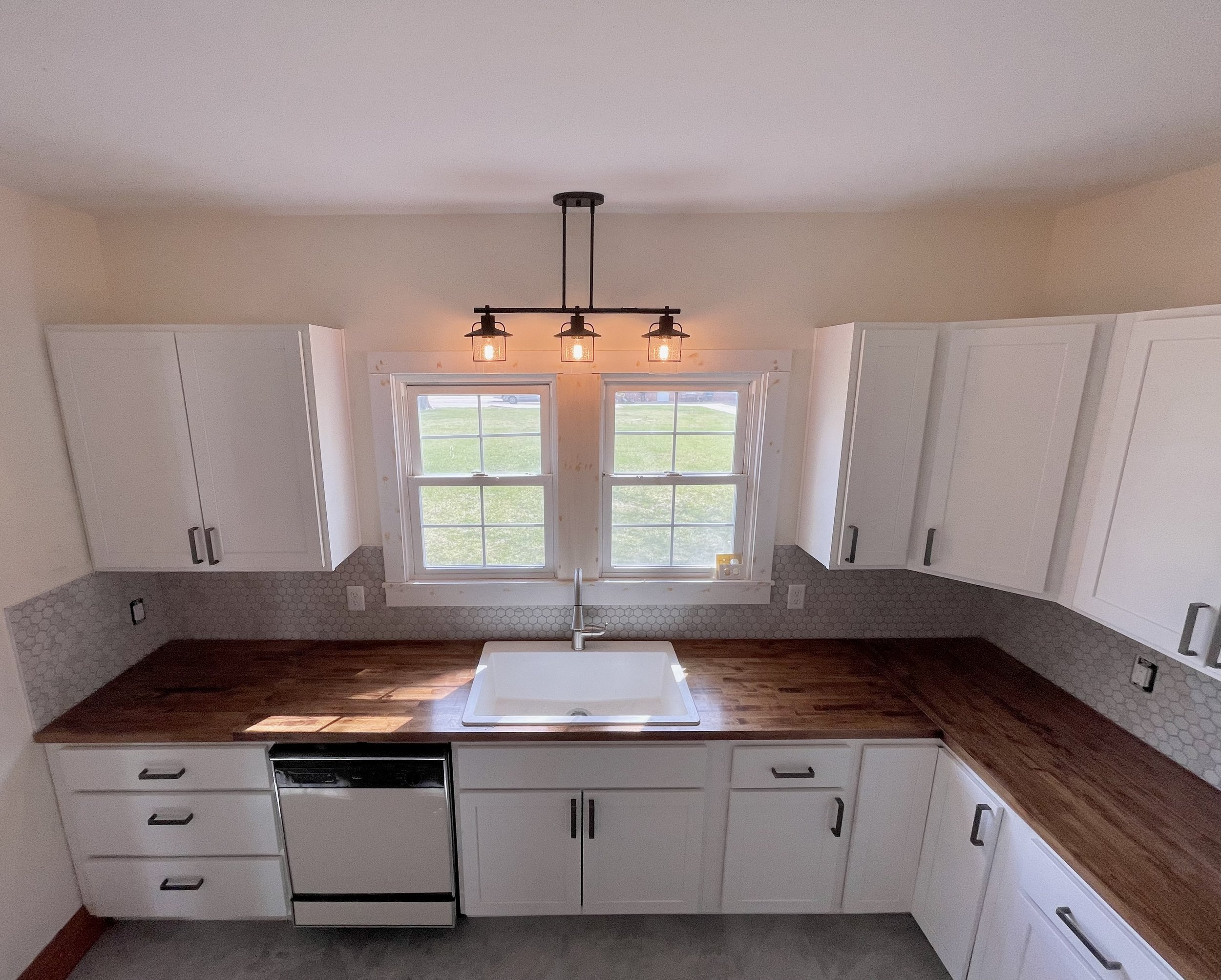 Kitchen with white cabinets, honeycomb tile backsplash, wooden countertop, a white sink under two windows, and a black industrial-style ceiling light fixture.