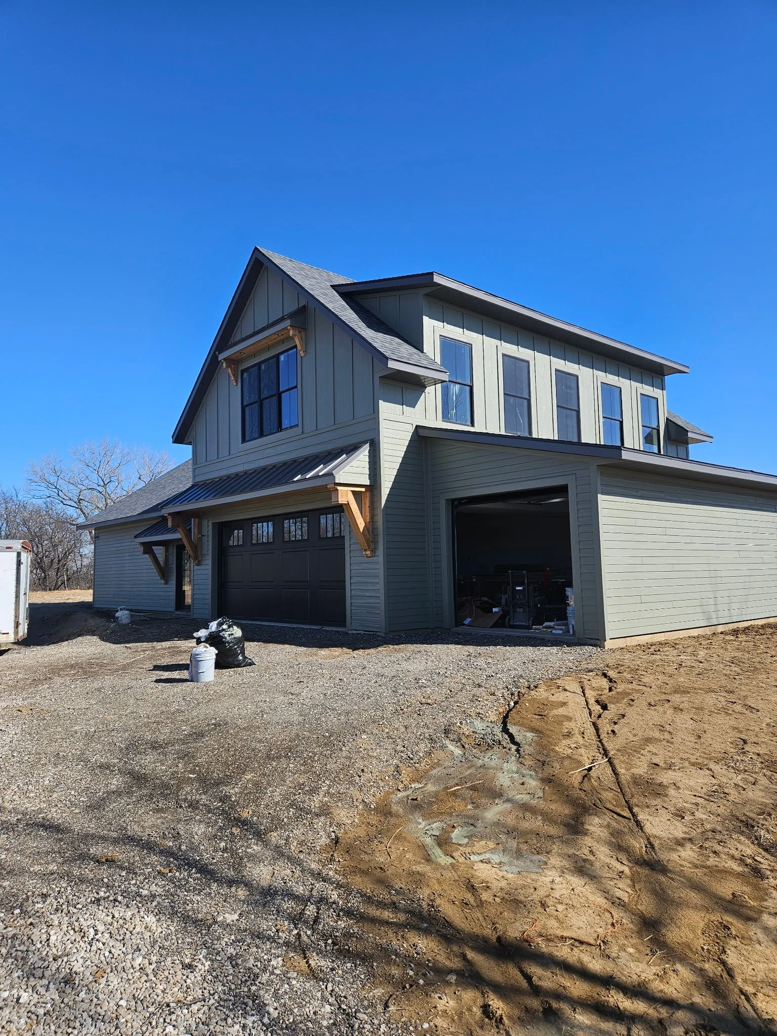 New two-story house under construction with dark garage door and siding, blue sky, and dirt yard.