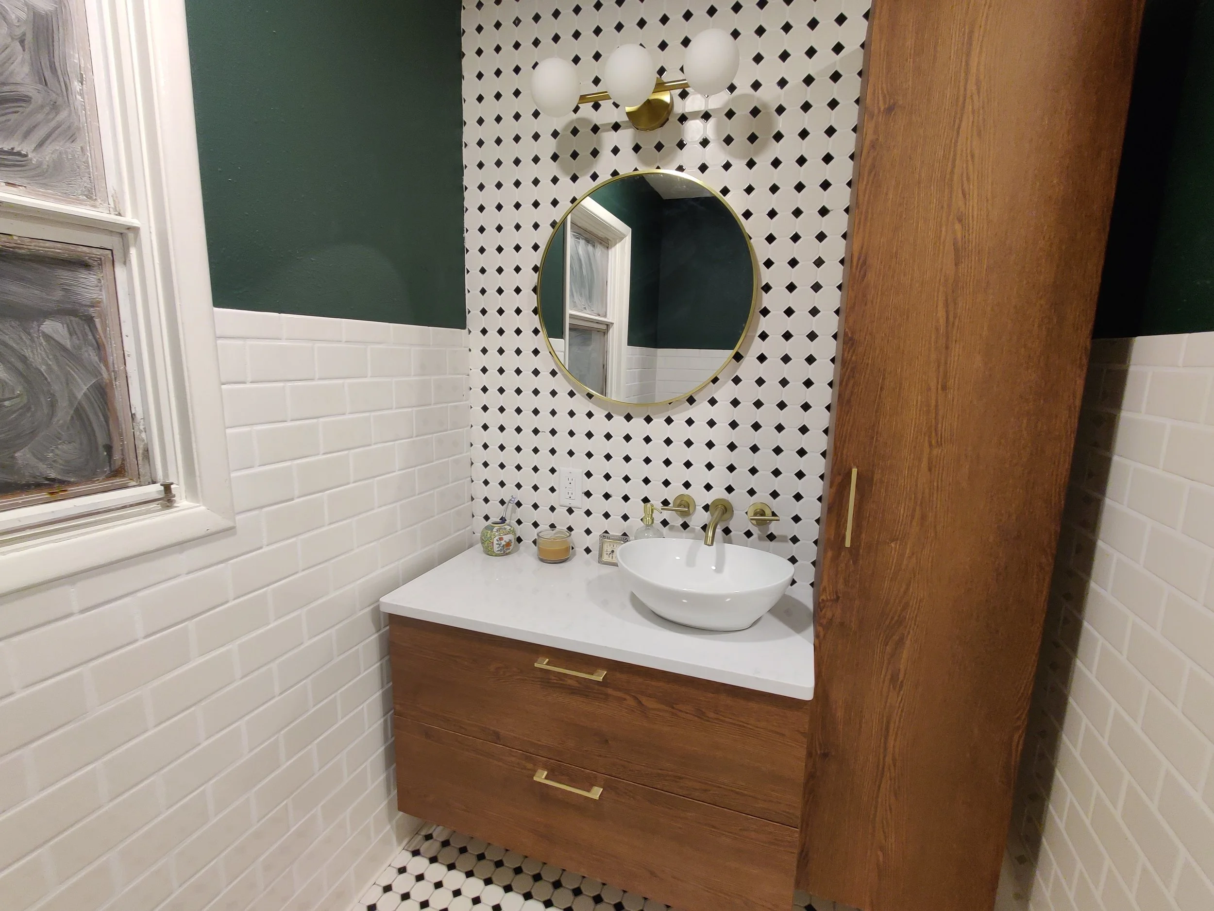 Modern bathroom vanity with white countertop, round vessel sink, gold fixtures, and a large wooden cabinet. Stylized black and white circular tile backsplash, dark green painted wall, and a small window with frosted glass in the background.
