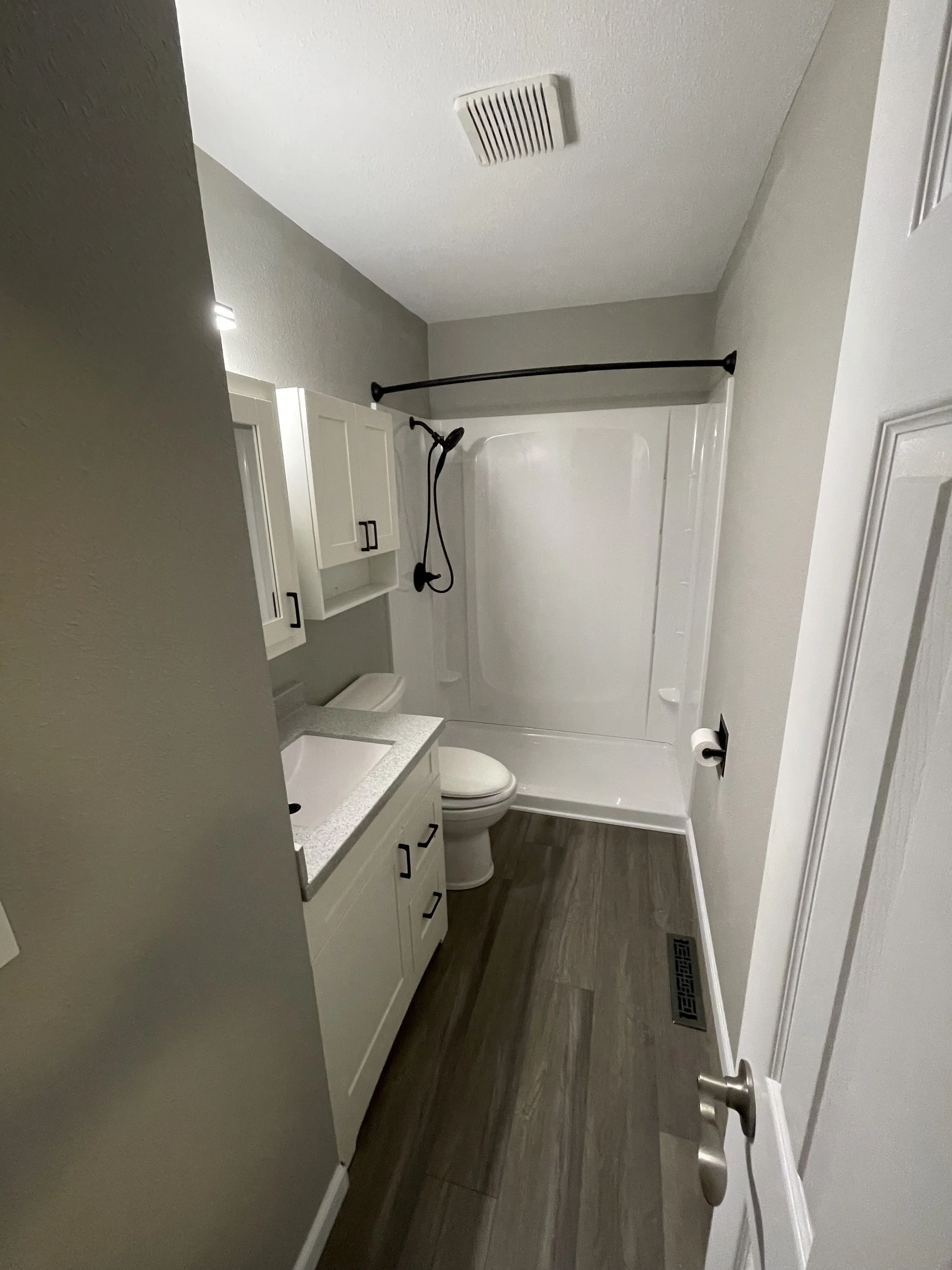 Small bathroom with white vanity, toilet, shower/tub combo with a black curtain rod, wood-look flooring, and light-colored walls.
