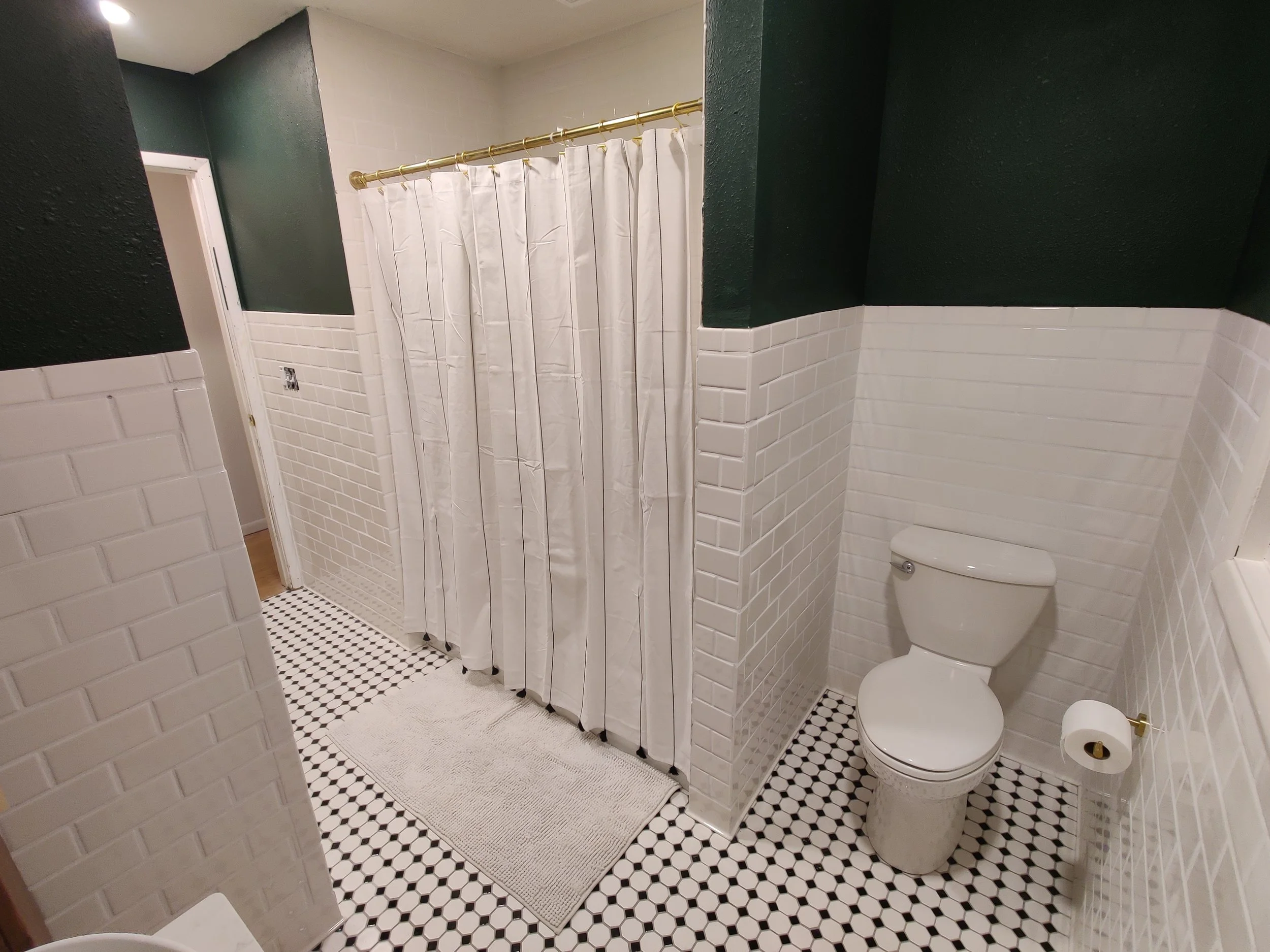 Bathroom with white brick walls, black and white hexagon floor tiles, a white toilet, a white shower curtain, a small white rug, and dark green upper walls.