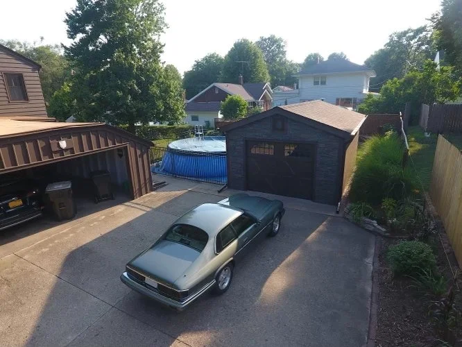 A car parked in a driveway next to a garage with an open door, a shed with a garage door, and a backyard with a pool, trees, and neighboring houses in the background.
