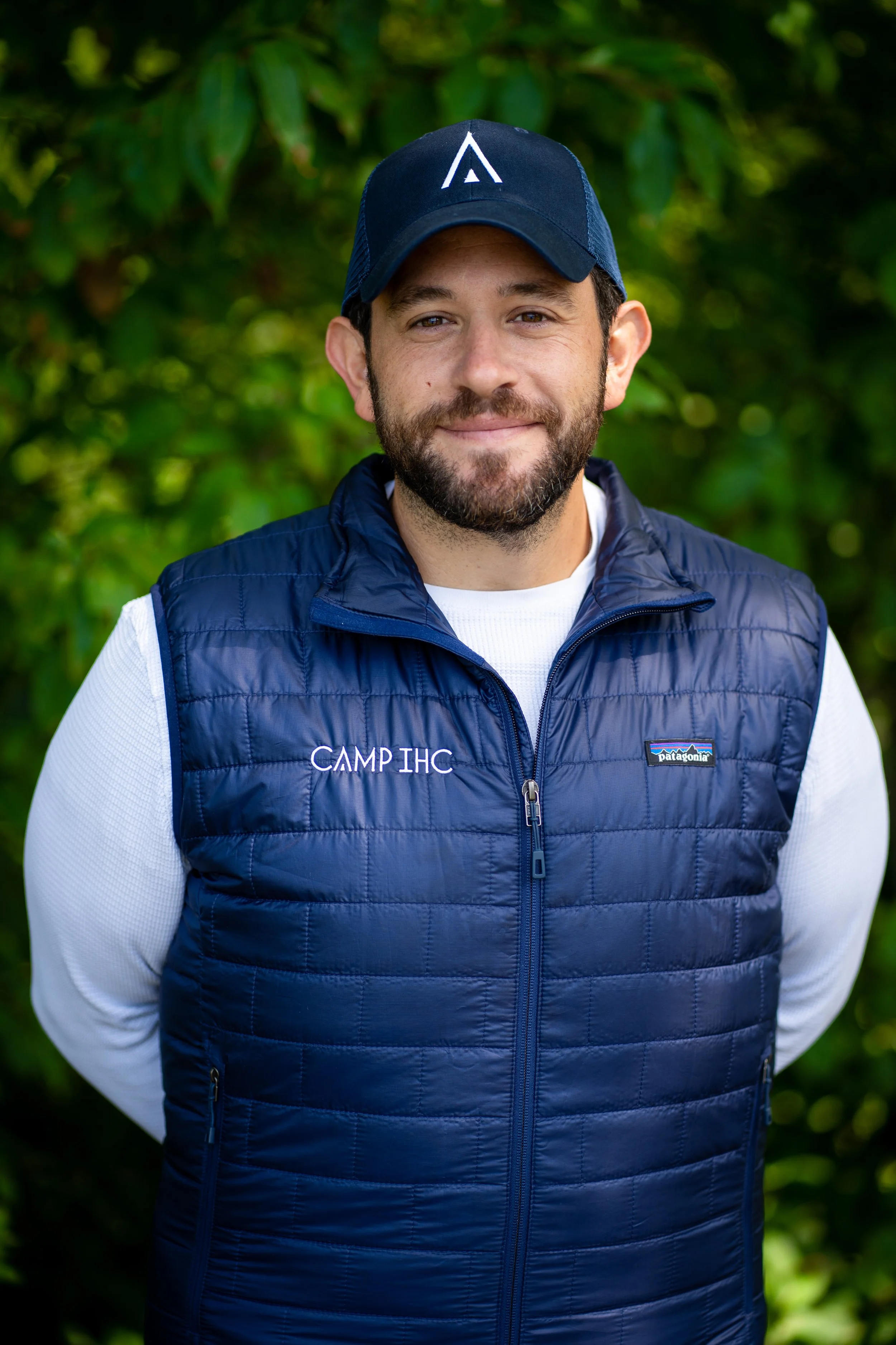 A man with a beard wearing a black cap, a navy blue quilted vest with the words "CAMP IHC" embroidered on it, and a white long-sleeve shirt, standing outdoors with green foliage in the background.