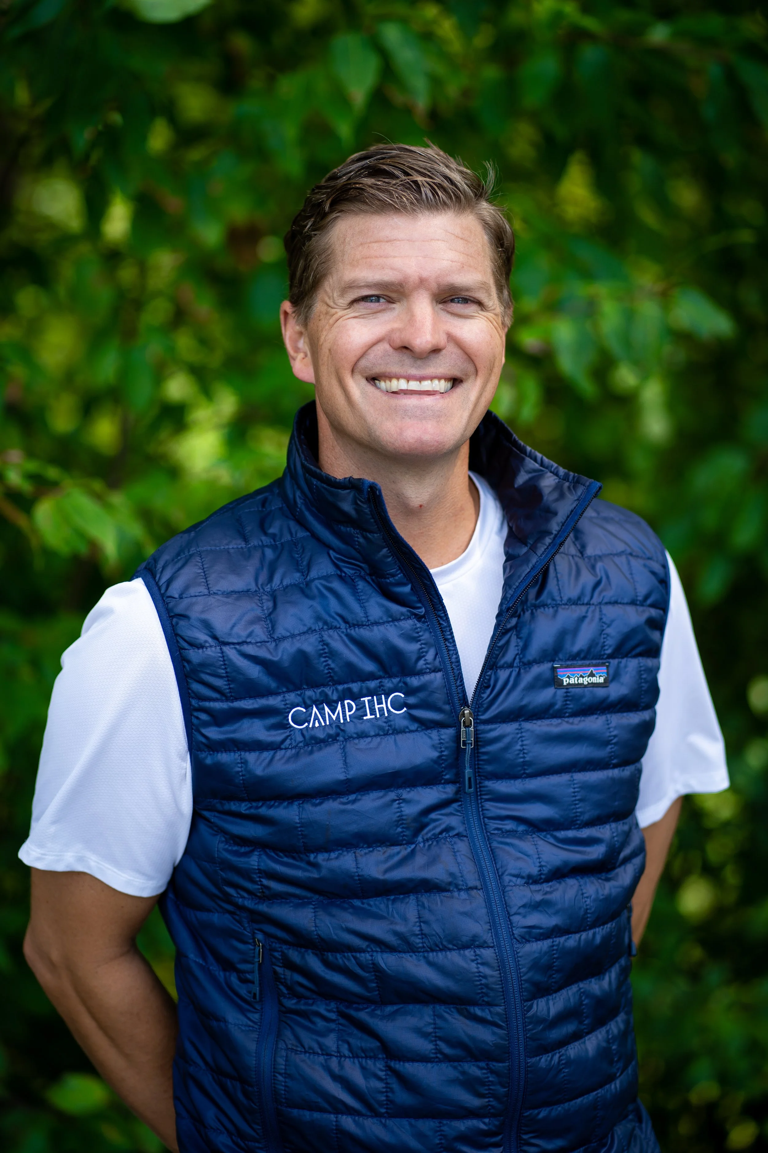 A smiling man wearing a white shirt and a blue Patagonia puffy vest with 'CAMP IHC' embroidered on it, standing outdoors with green foliage in the background.