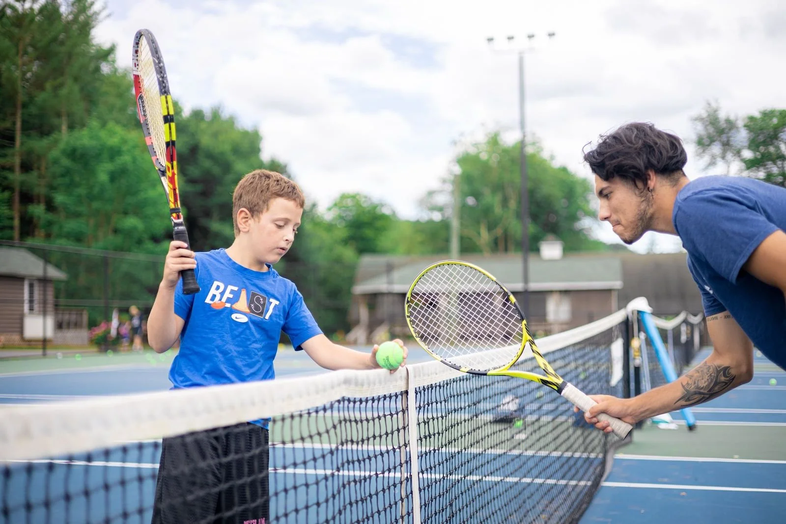 A young boy wearing a blue Nike t-shirt holding a tennis racket and a tennis ball balanced on the net while an adult man with a tattoo on his arm holding a tennis racket leans over to teach him on a tennis court outdoors.
