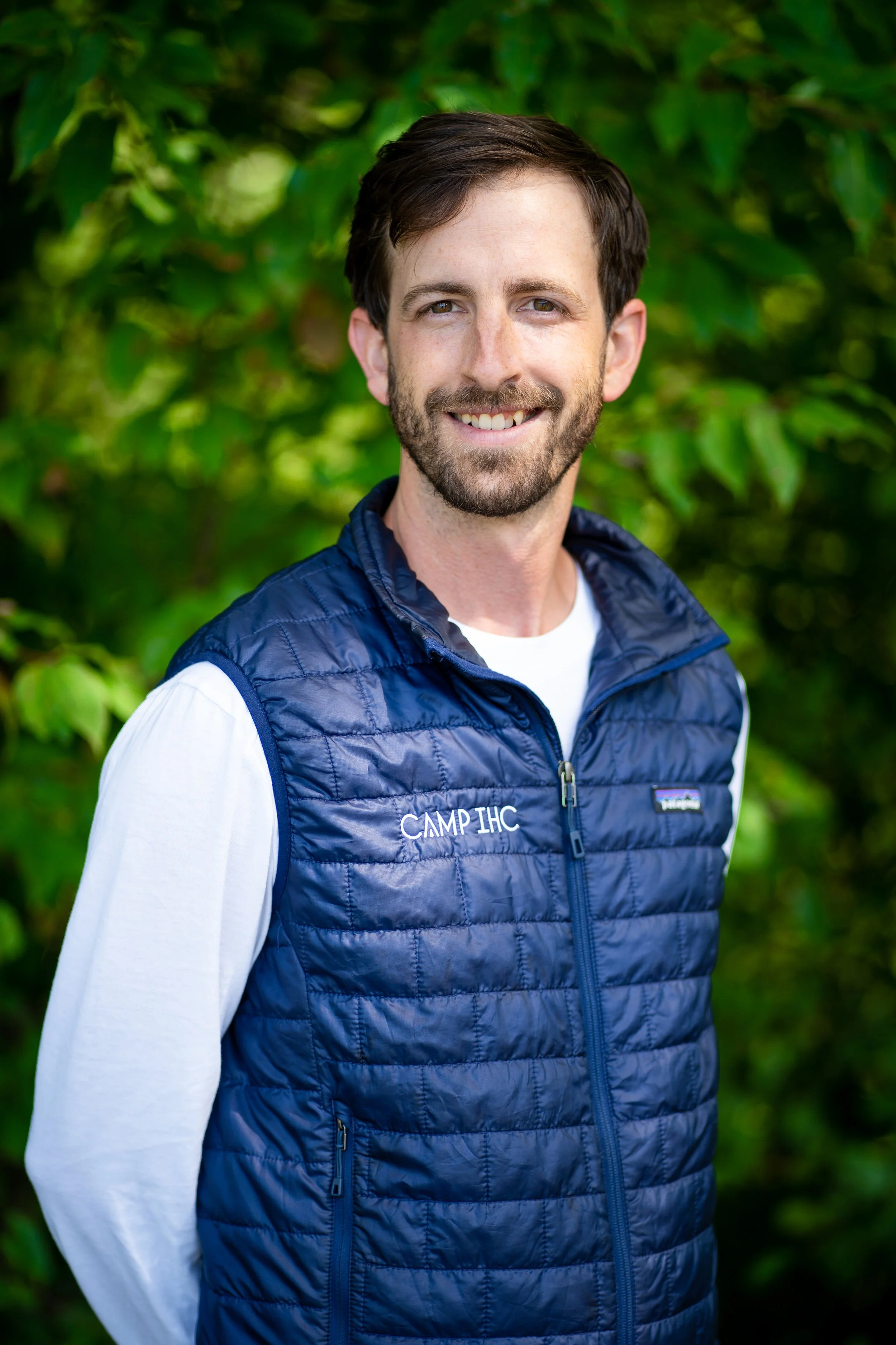 A man with brown hair and a beard smiling outdoors, wearing a navy blue puffer vest with 'CAMP IHC' embroidered on it, over a white long-sleeve shirt, with green foliage in the background.
