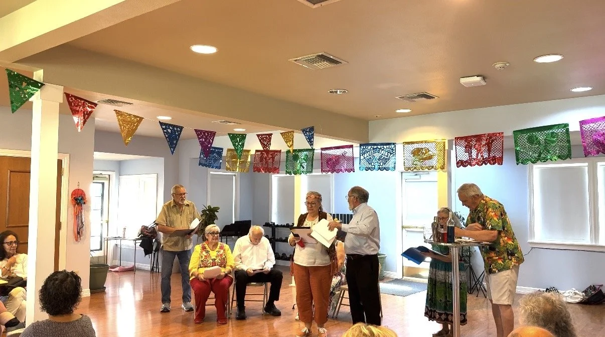A group of older adults gathered in a decorated room with colorful banners, participating in a social or community event. Some are standing and reading from papers, while others are seated.