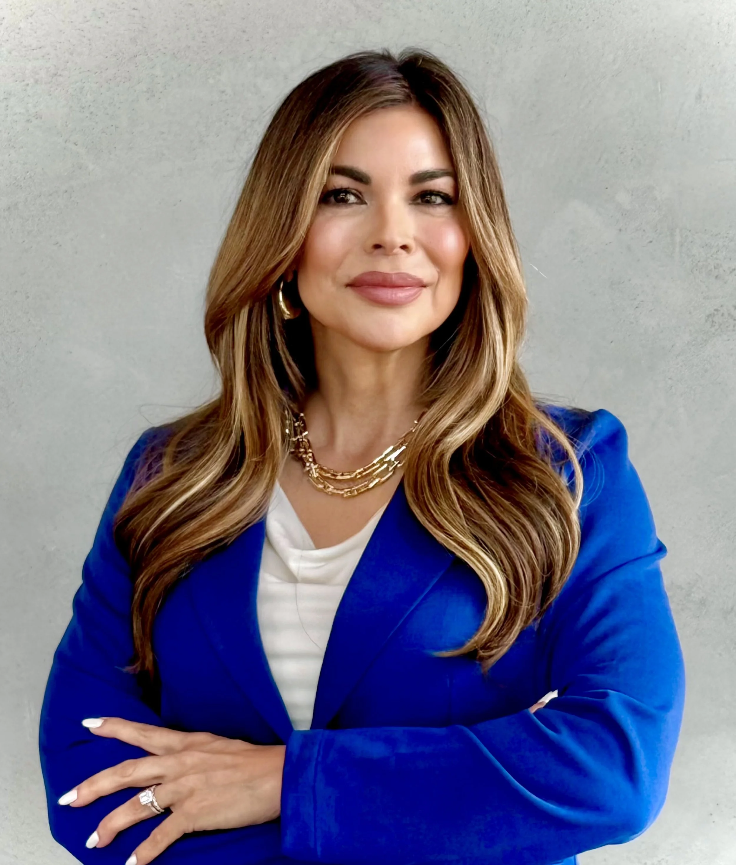Woman with long chestnut hair and light makeup wearing a royal blue blazer, layered gold necklaces, and a white top, smiling with arms crossed in front of a plain light gray background.