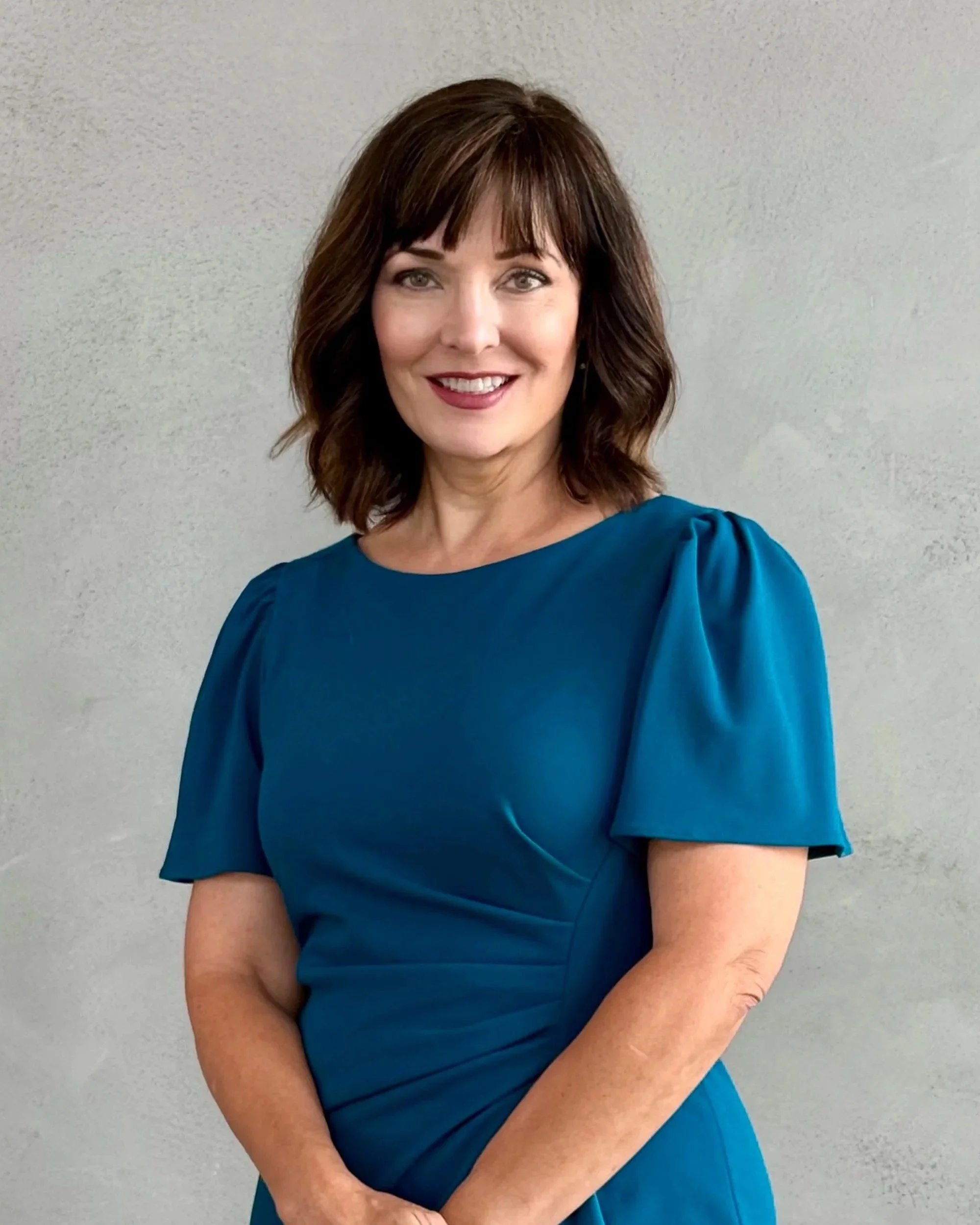A woman with short, dark brown hair and bangs, smiling, wearing a blue dress with puffed sleeves, standing against a light gray wall.