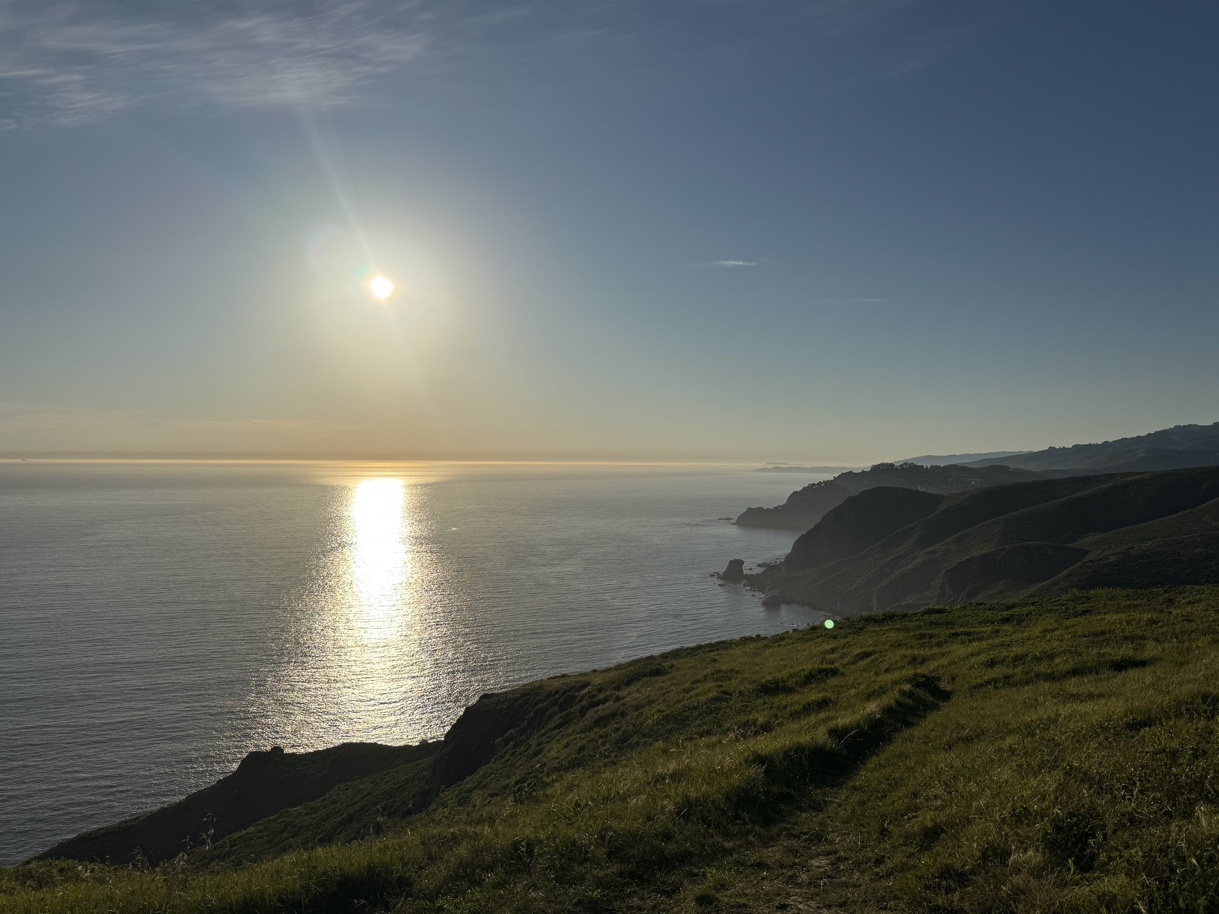 Golden sunset over the Pacific Ocean from a grassy coastal cliff, with sunlight reflecting on the water and rugged shoreline stretching into the distance along the California coast.