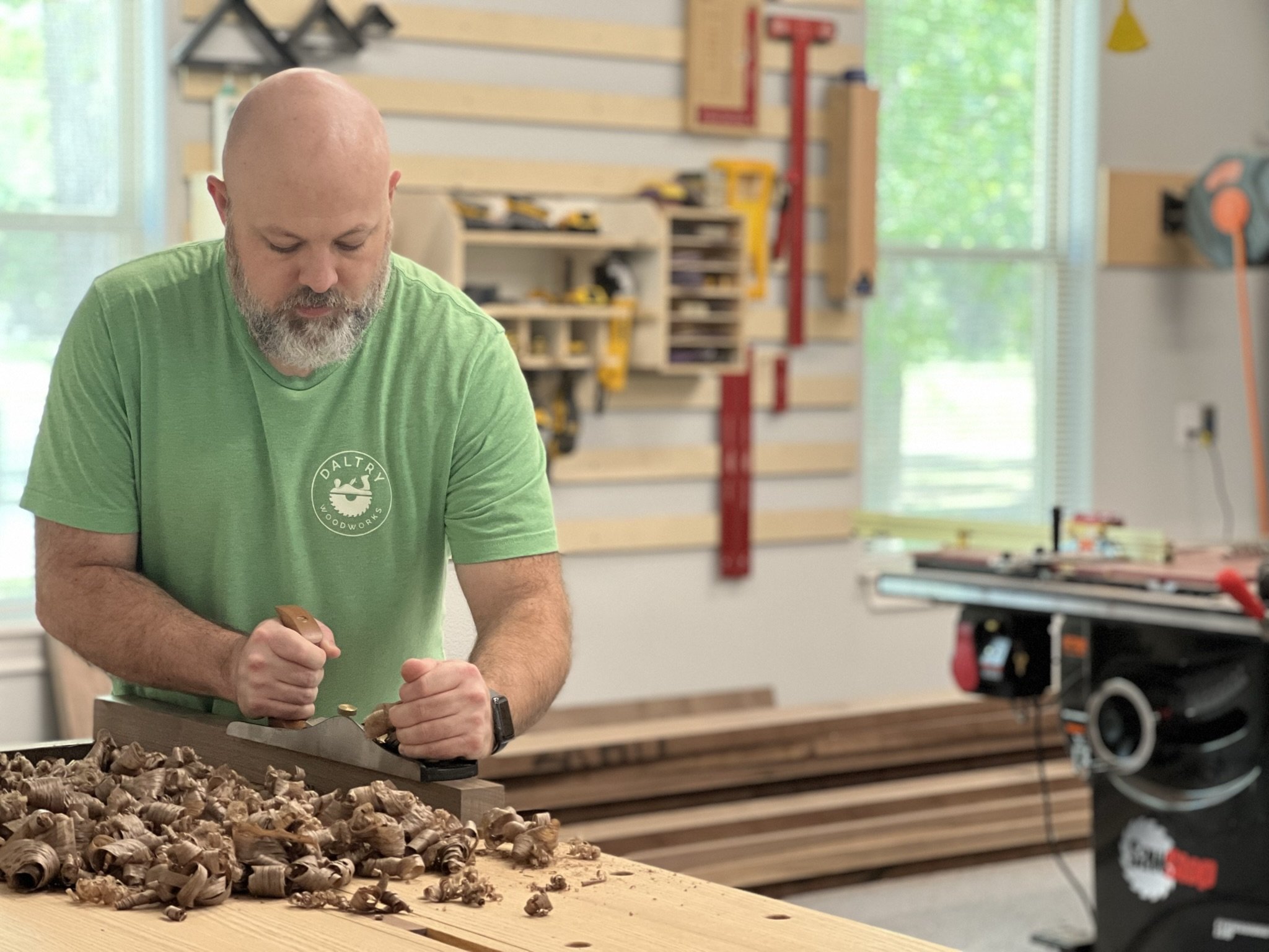 A man using a hand plane to smooth wood in a workshop. He is wearing a green shirt, and there are tools and equipment in the background.