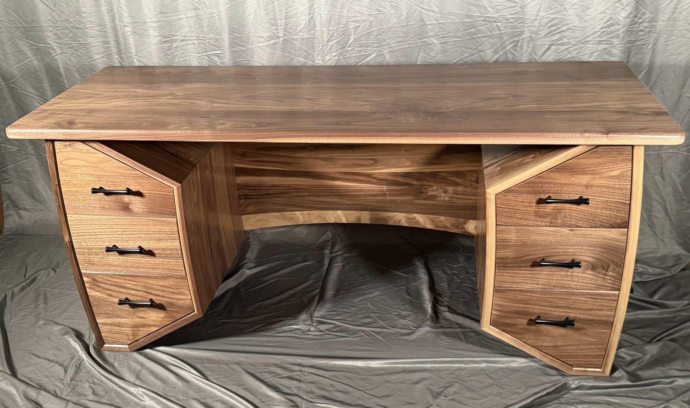 Wooden desk with natural finish, featuring six drawers with black handles, on a gray fabric backdrop.