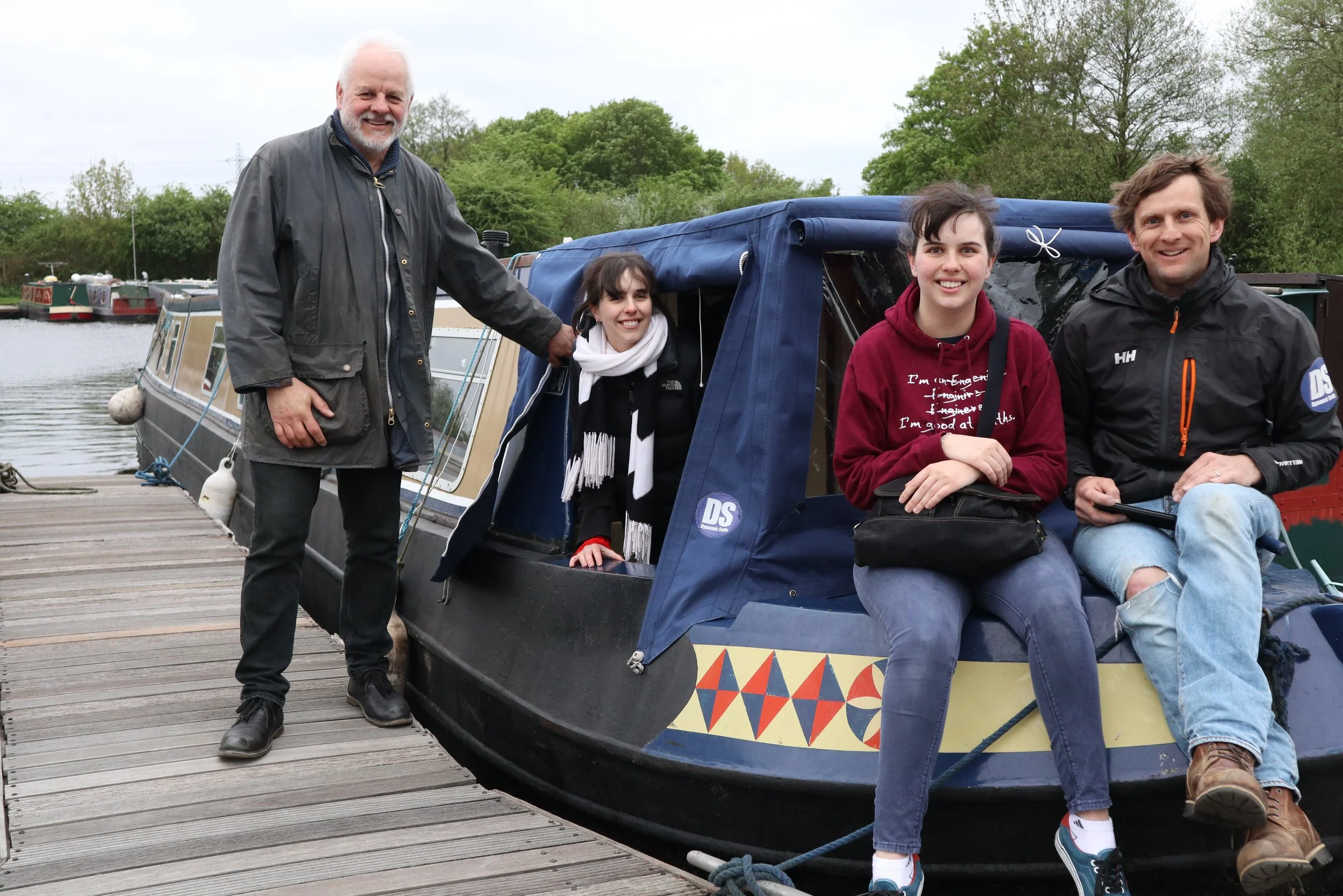 Four happy people at the dock with a narrowboat, greenery, and trees in the background. One man standing on the dock holding the boat, three people sitting on the boat.