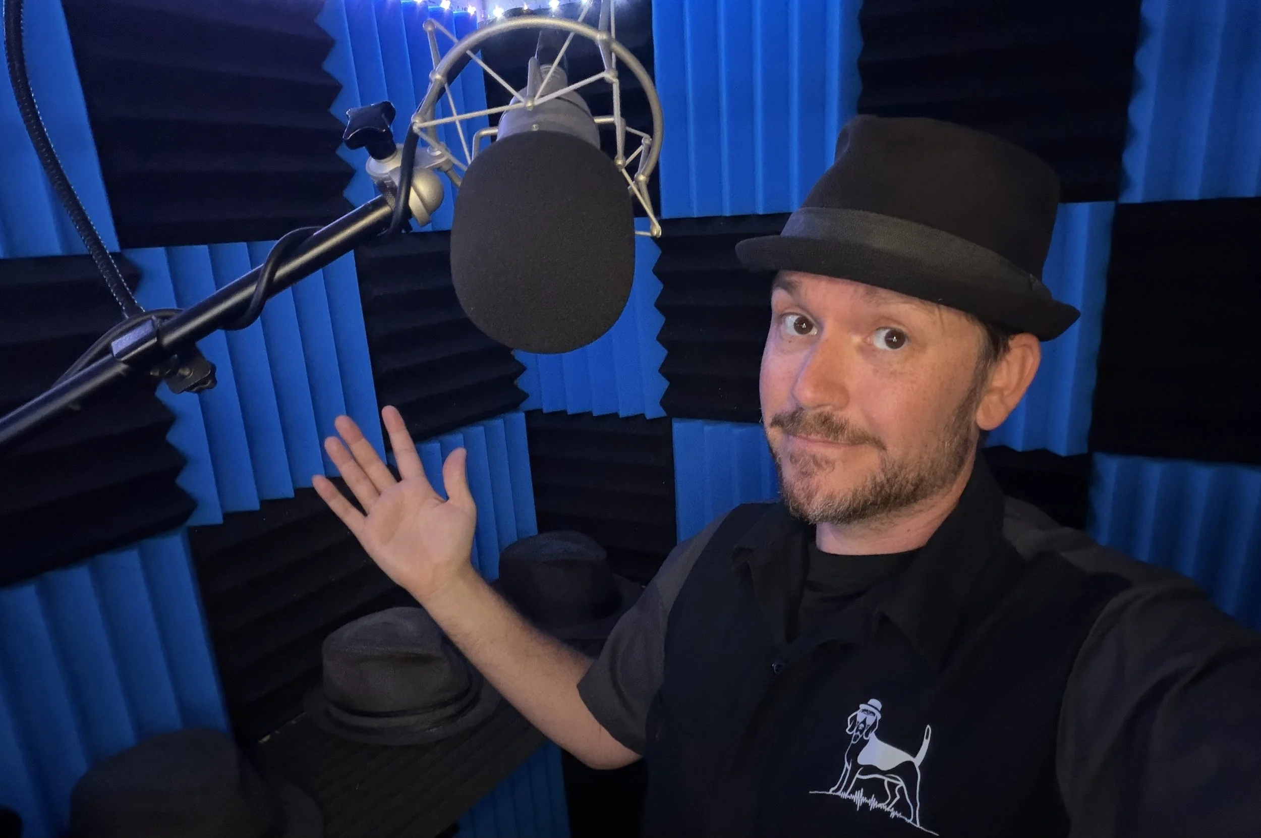 Orlando-based voice actor, audio producer, an performance coach Nate of Begle Booth Studios gesturing toward a Neumann TLM 103 microphone and assortment of hats inside is professional recording studio, surrounded by acoustic foam treatment