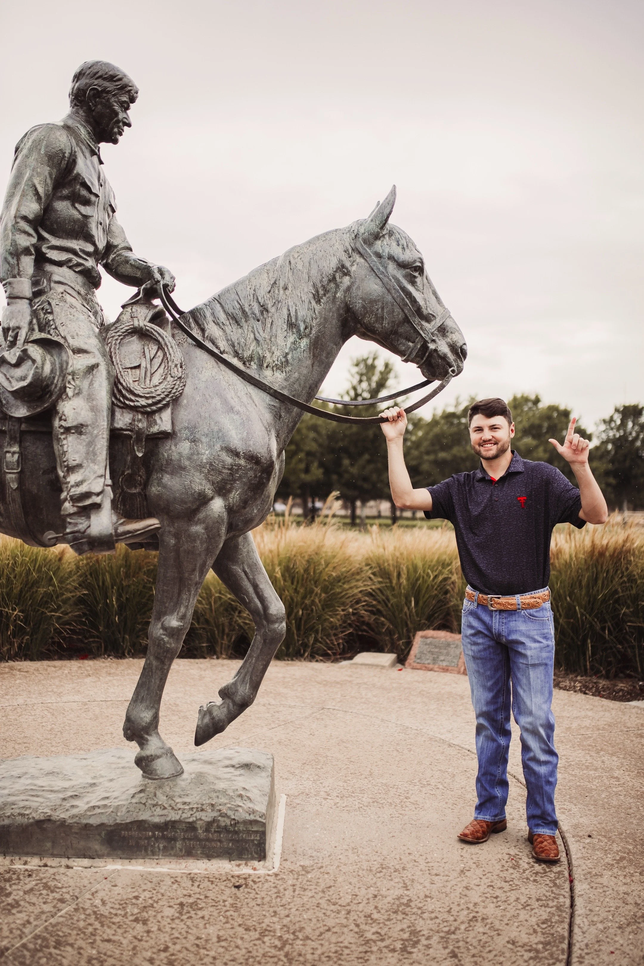 A young man standing next to and posing with a statue of a mounted soldier, waving one hand and pointing up with the other, outdoors on a paved area with grass and trees in the background.