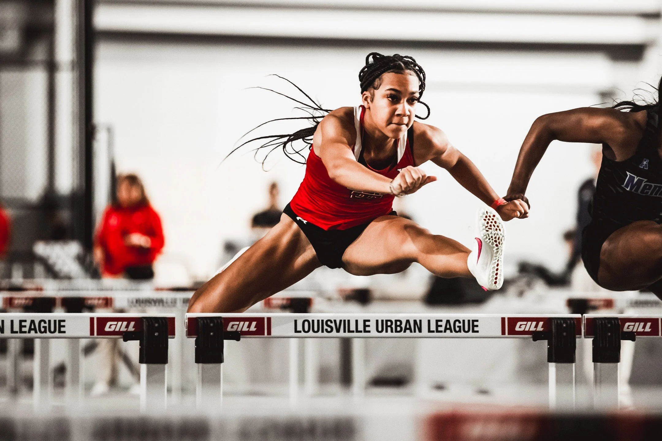Female athlete in red uniform competing in a hurdles race at the Louisville Urban League track and field meet.