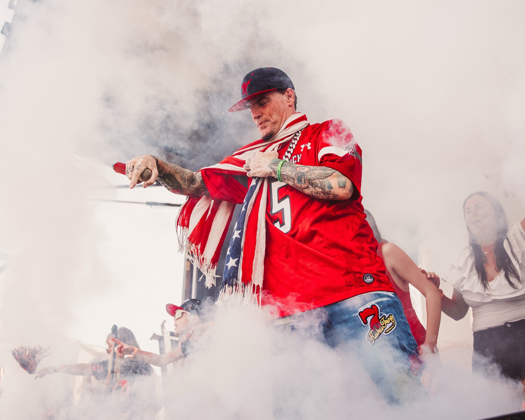 Man wearing a red sports jersey, tattoos, and a baseball cap, surrounded by smoke, holding a red flare, with others in the background.