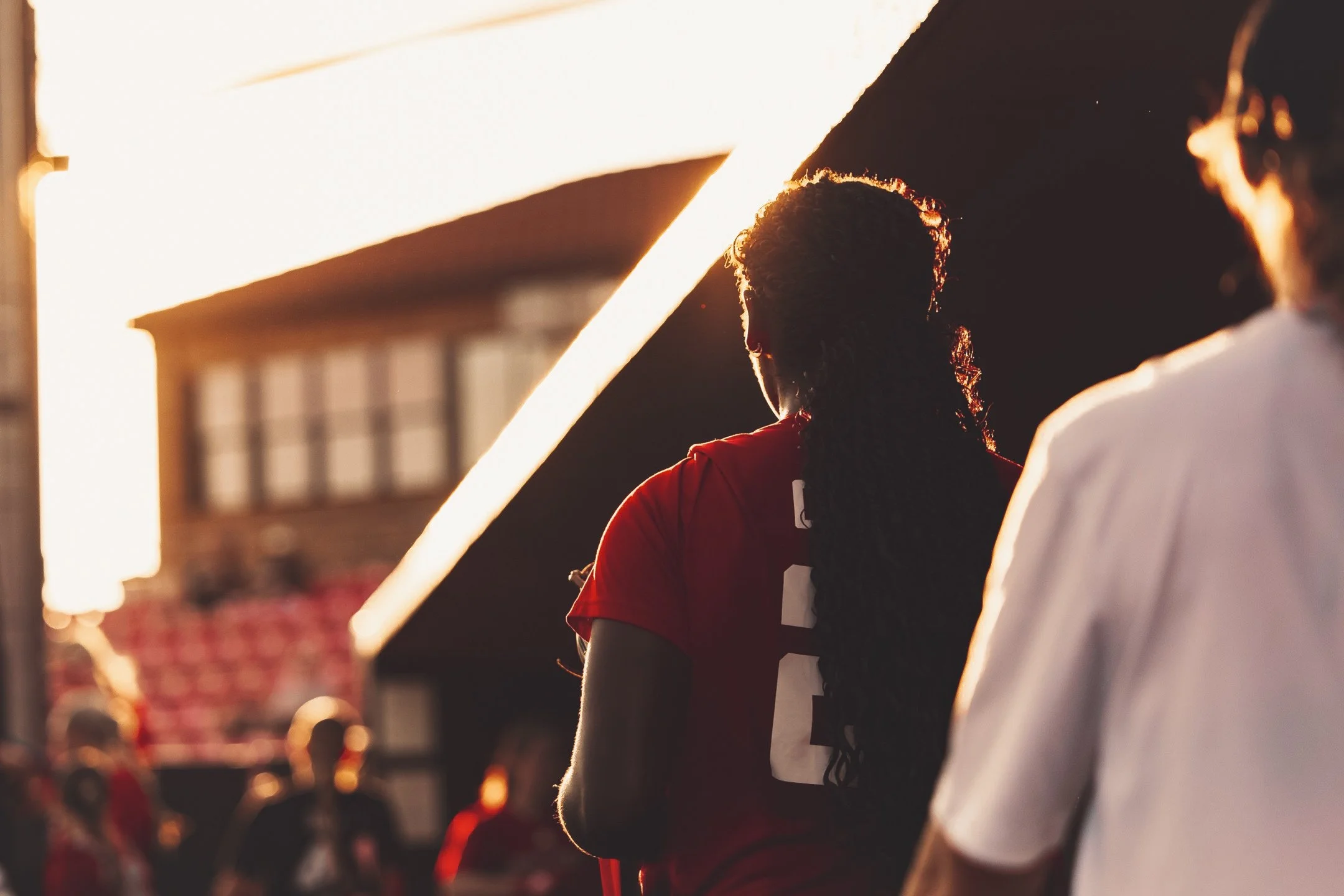 People standing outdoors during sunset, silhouetted against the light, with a building in the background.