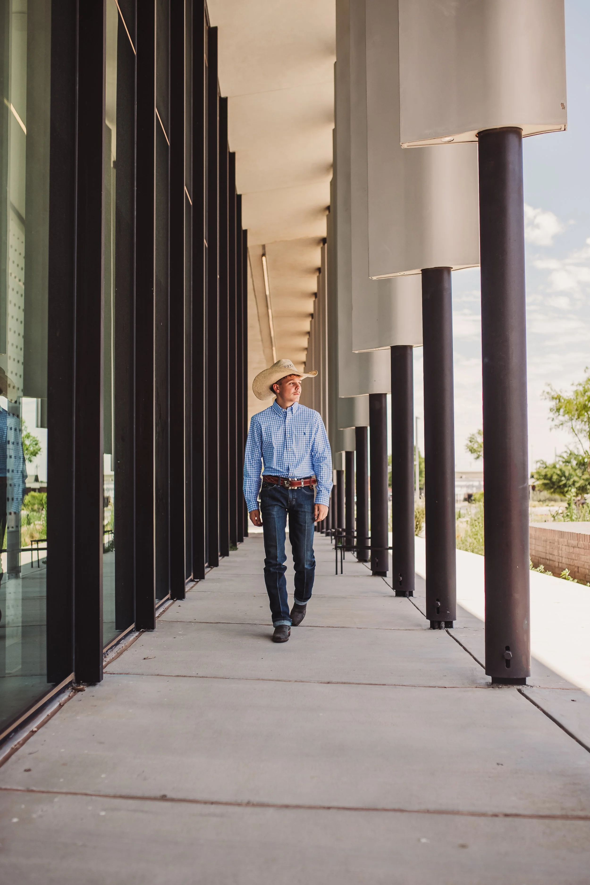 A young man dressed in cowboy attire, including a straw cowboy hat, blue checkered shirt, and jeans, walking along a covered sidewalk outside a modern building with tall glass windows and dark support columns.
