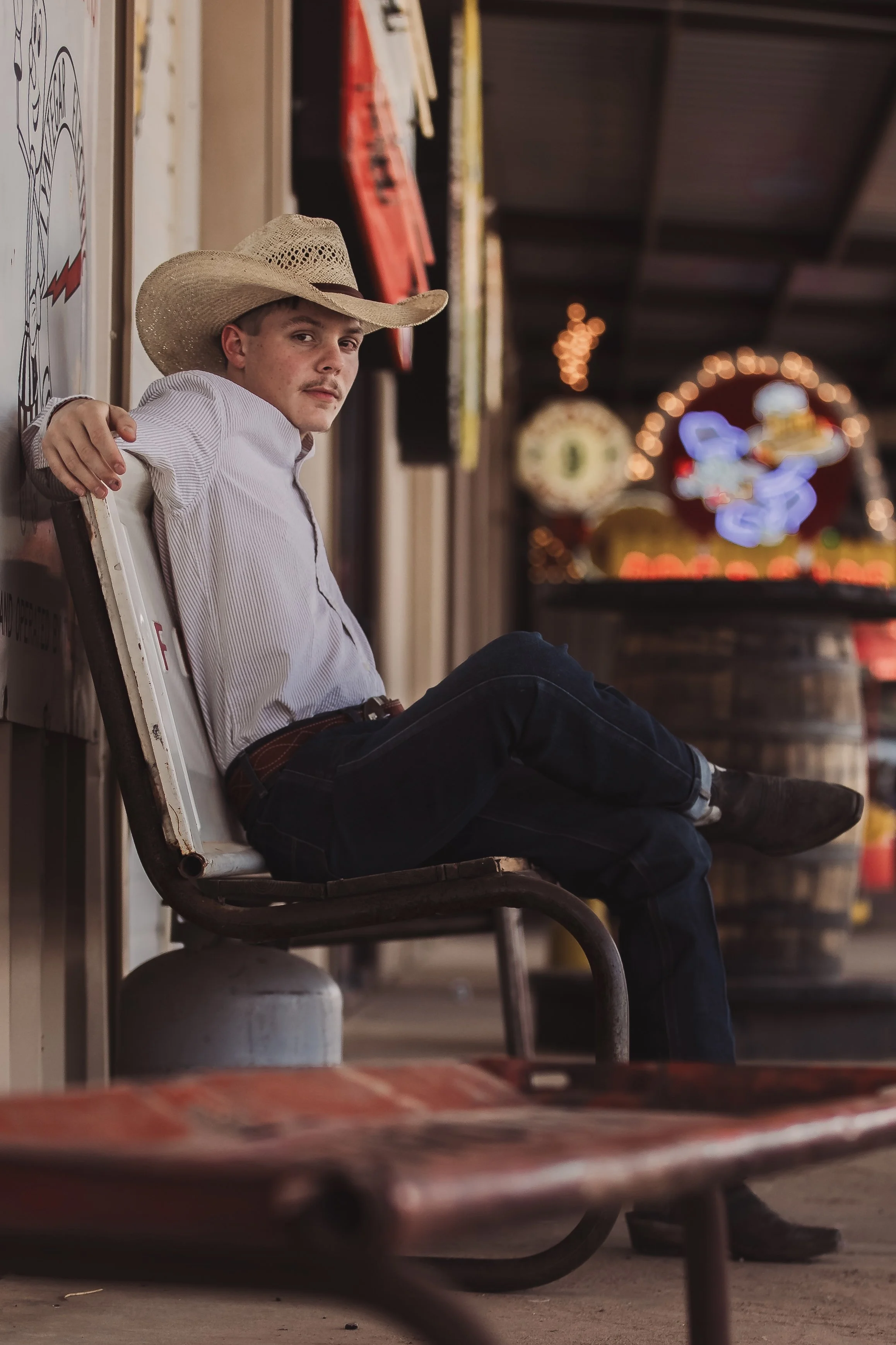 A man sitting on a bench outside a building. He is wearing a cowboy hat, light-colored shirt, dark pants, and boots. The background features blurred colorful lights and signs.