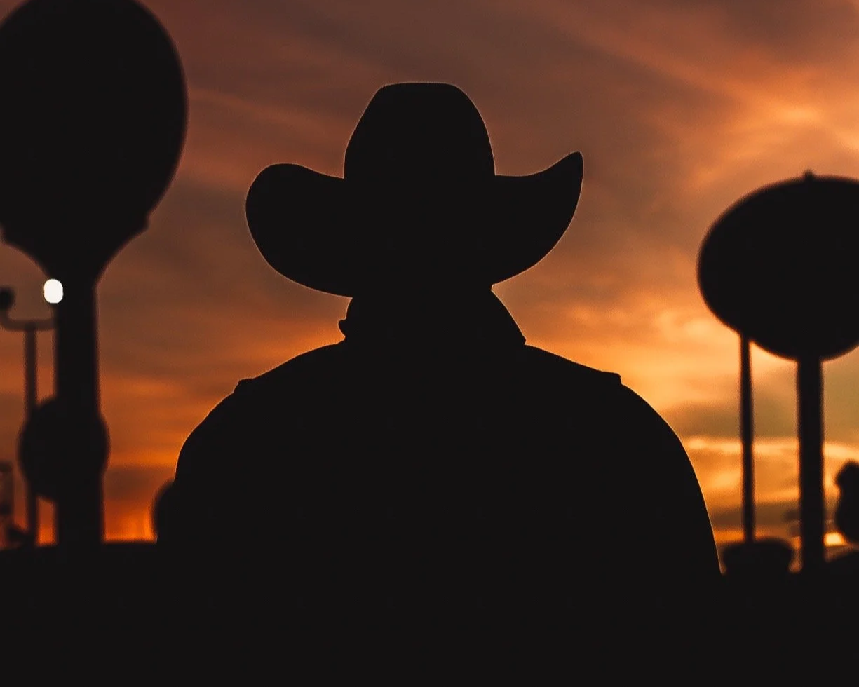 Silhouette of a person wearing a cowboy hat against a sunset sky with a few oil pump jacks in the background.