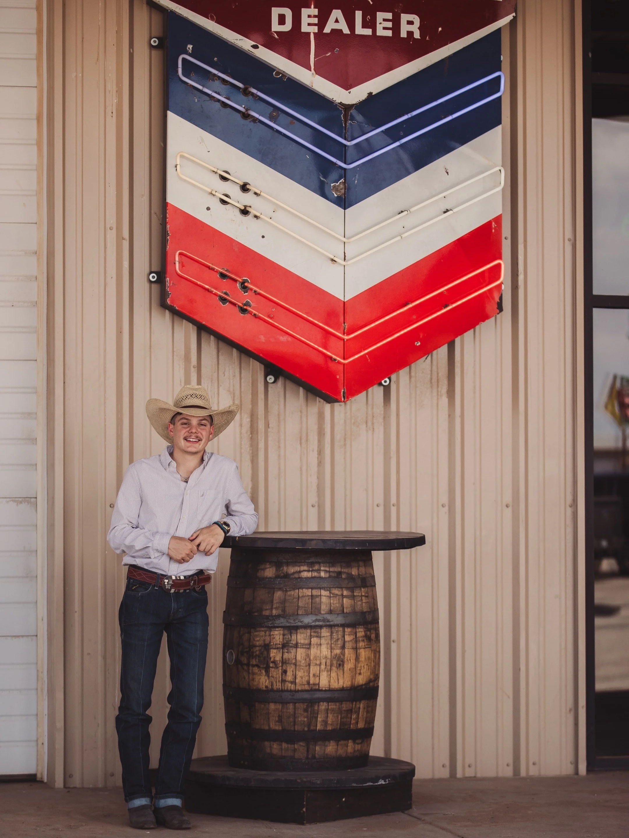 A young man wearing a cowboy hat, white shirt, and jeans standing next to a round wooden table outside a building with a striped metal wall and neon sign that reads 'DEALER' in red, white, blue, and purple colors.