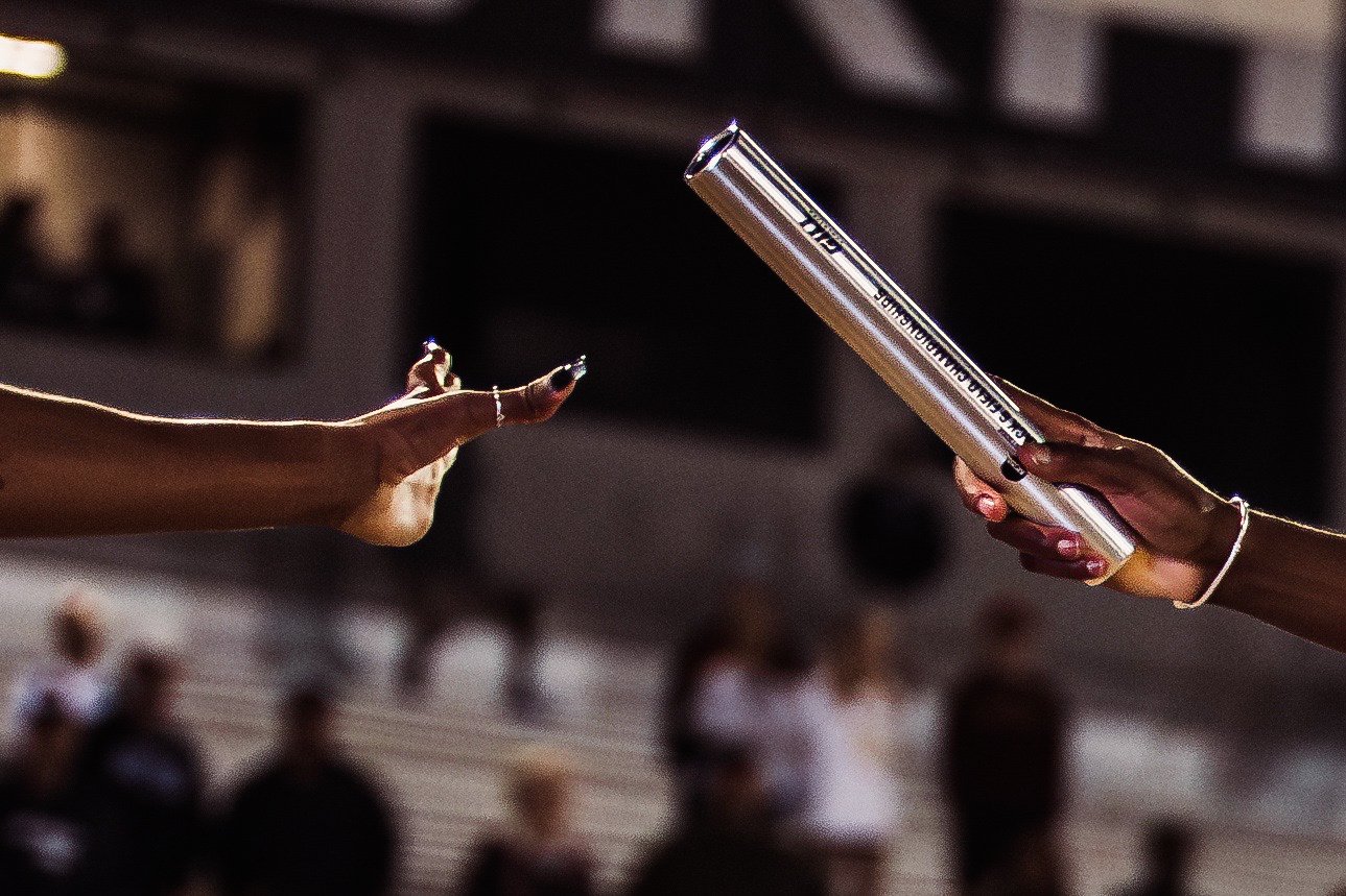 Two hands exchanging a rolled-up diploma or certificate in a festive or formal setting, with an audience in the background.