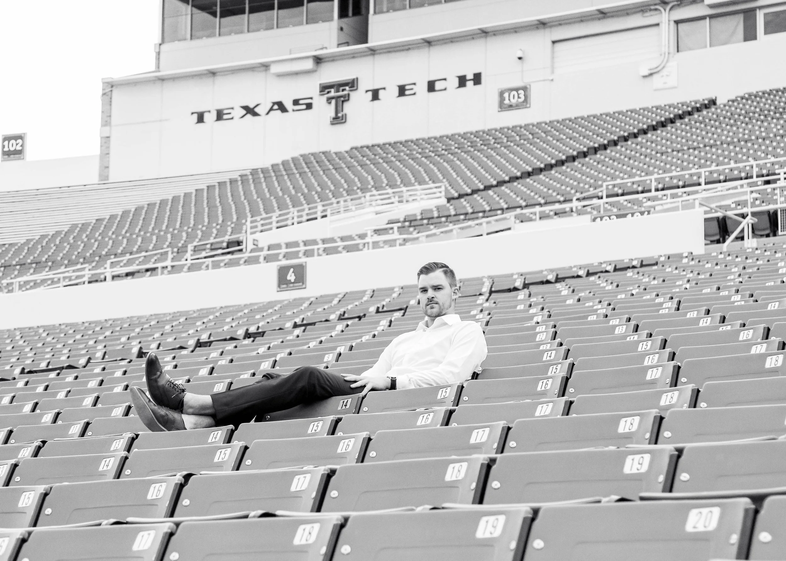 A man in a white dress shirt and black pants sitting alone on a stadium seat at Texas Tech stadium with empty seats around him.