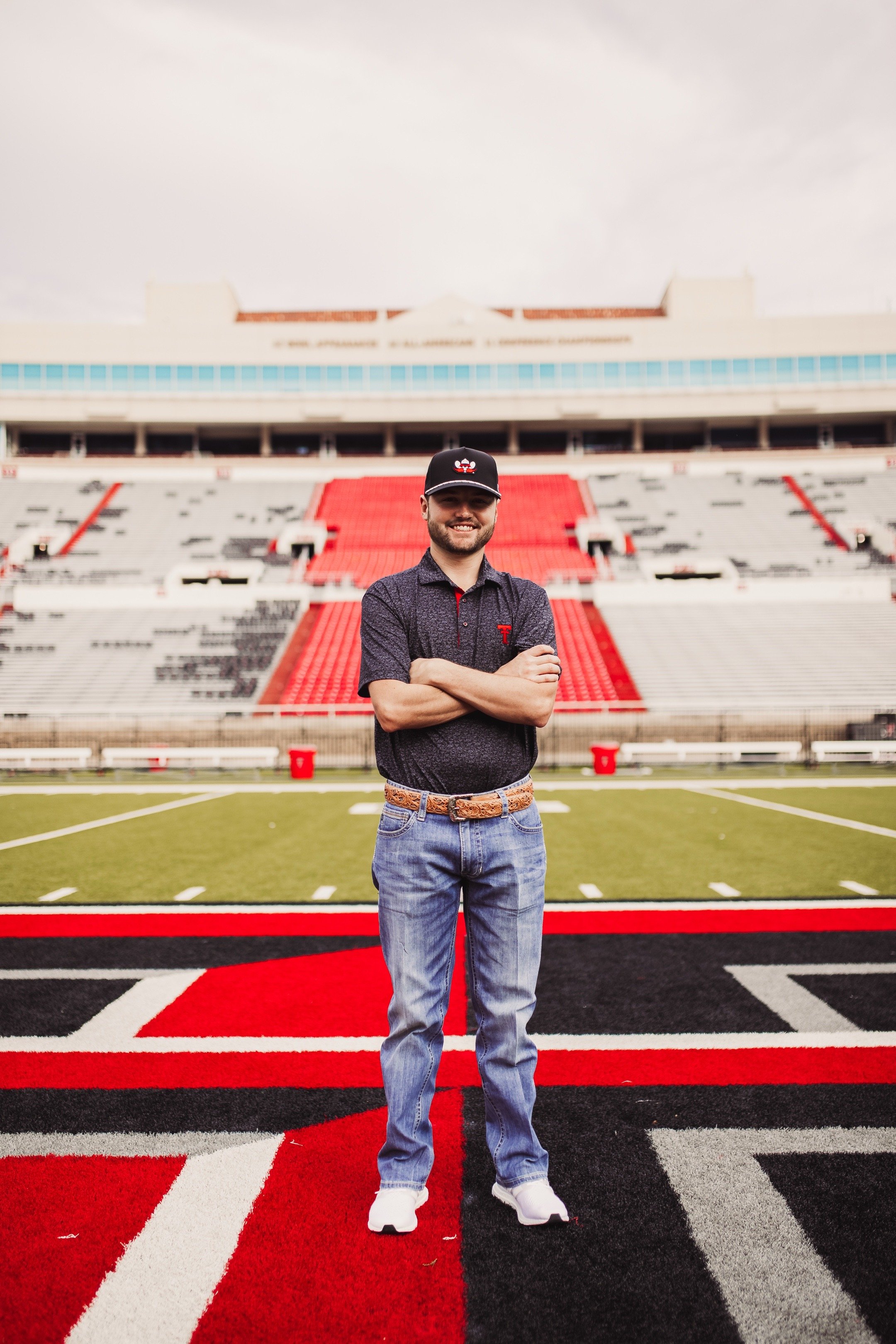 A man standing on a sports field with arms crossed, smiling, wearing a black baseball cap, a dark polo shirt, jeans, and white sneakers, in an empty stadium.