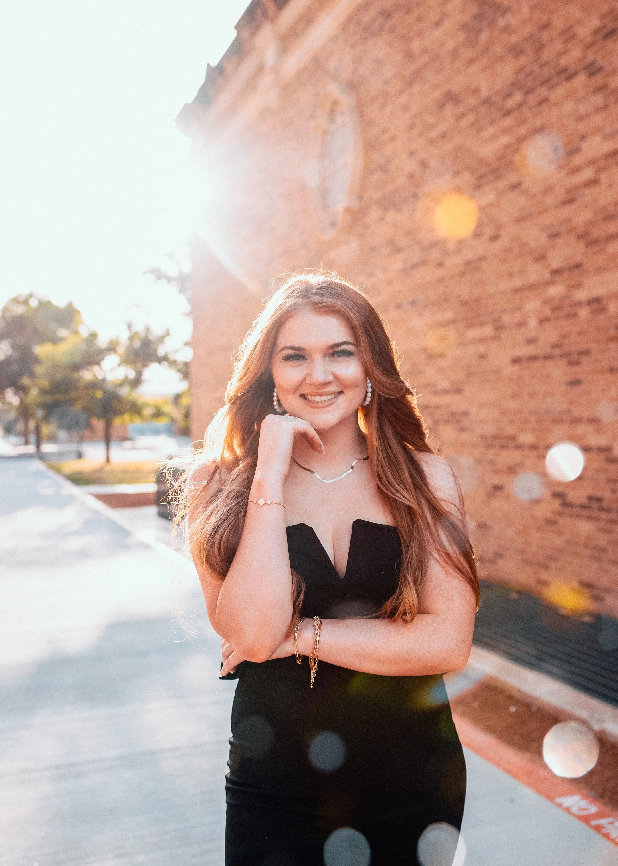 A young woman with long red hair in a black dress standing outside in sunlight, smiling, with a brick wall and trees in the background.
