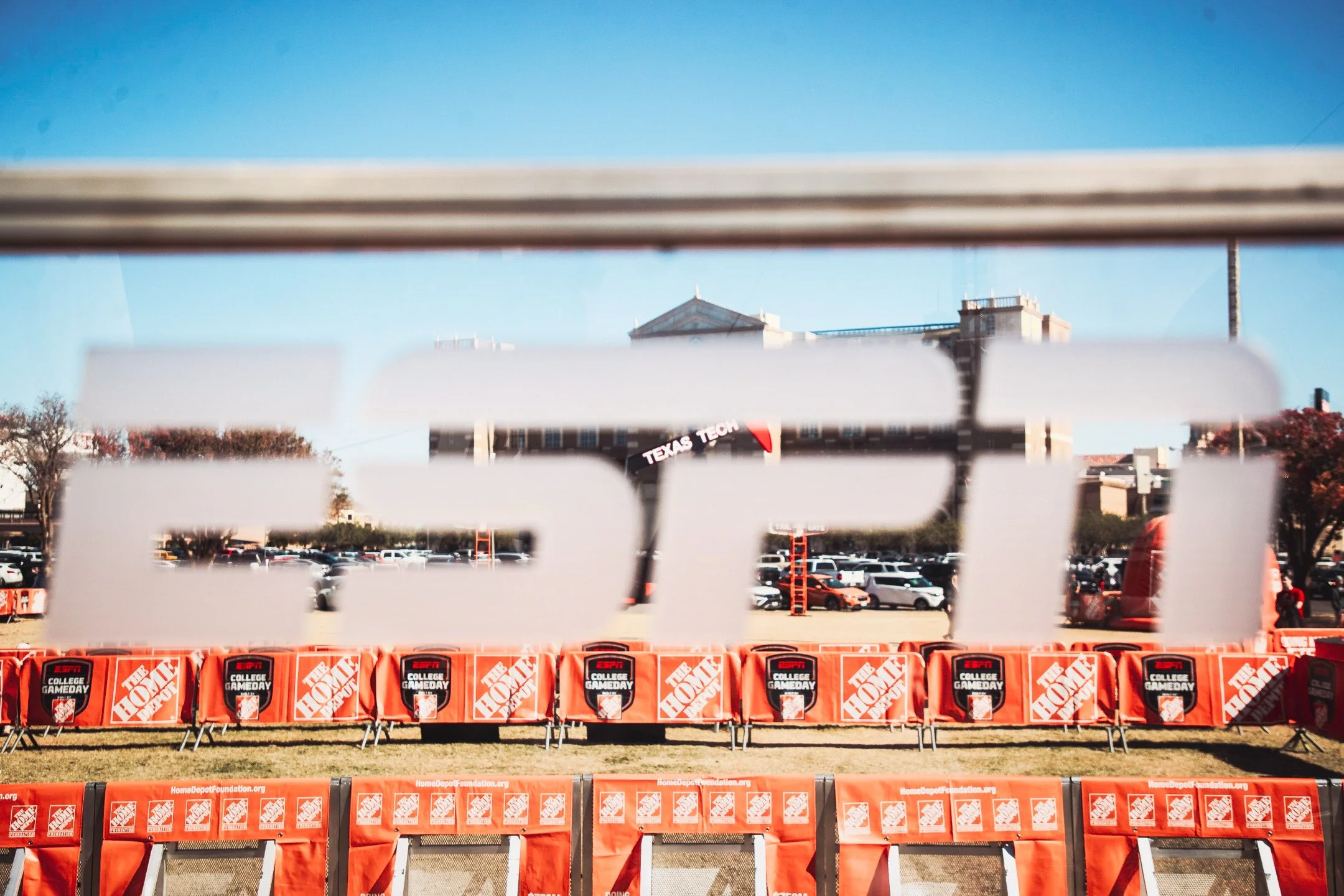 Photo taken through a window with the ESPN logo covering the view of an outdoor event area, which is decorated with orange barriers displaying the Home Depot logo and the words 'College Gameday'. In the background, there are people, trees, cars, and 
