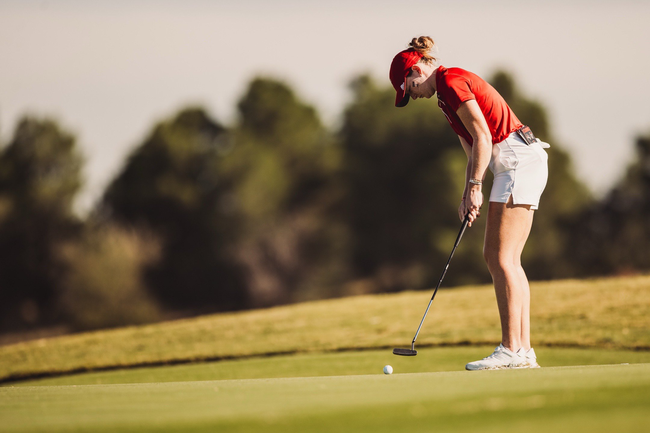 A woman dressed in a red shirt, white shorts, and a red cap preparing to putt in golf on a grassy course with trees in the background.