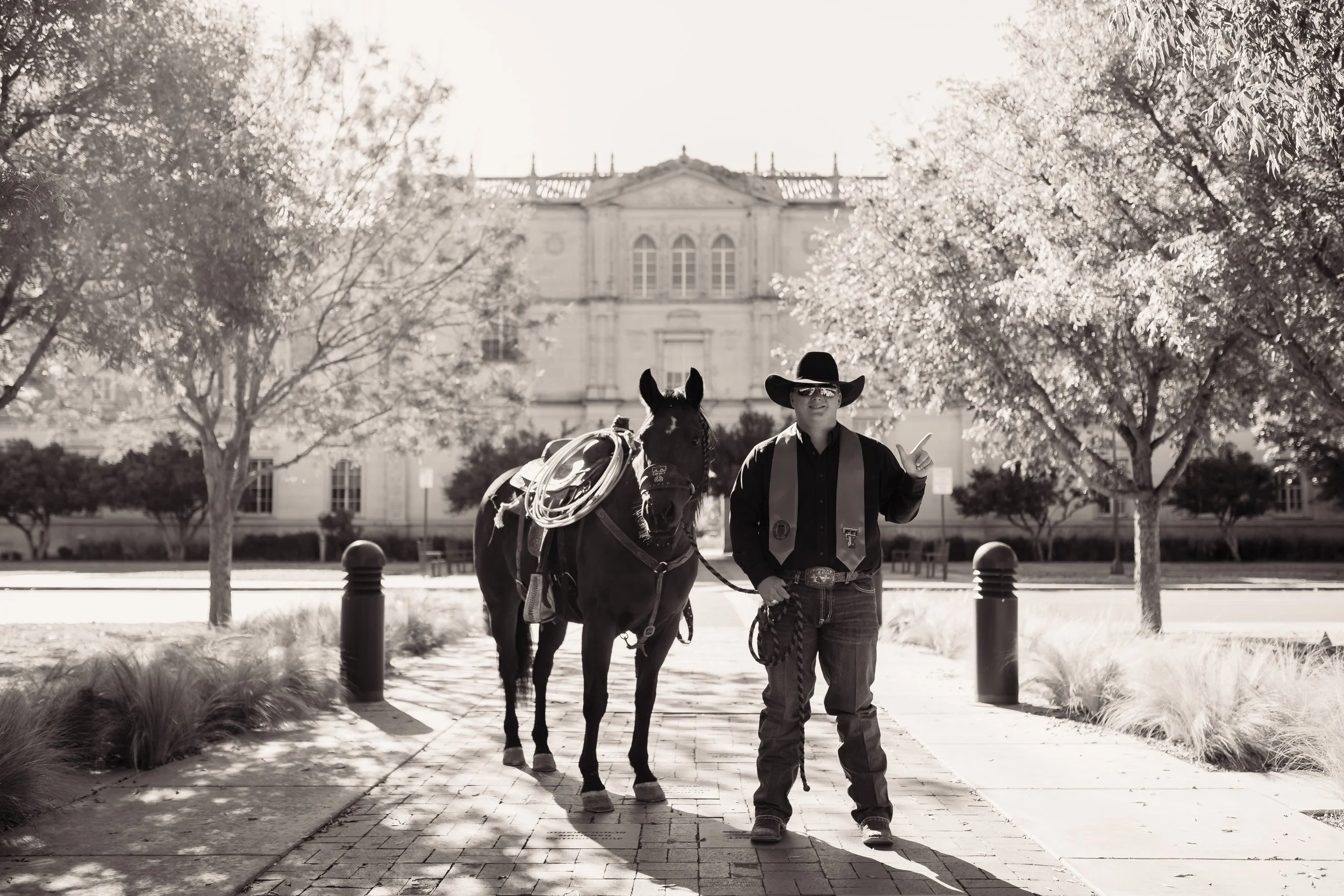 A man wearing a cowboy hat, sunglasses, and western clothes walking a horse along a sidewalk lined with trees.