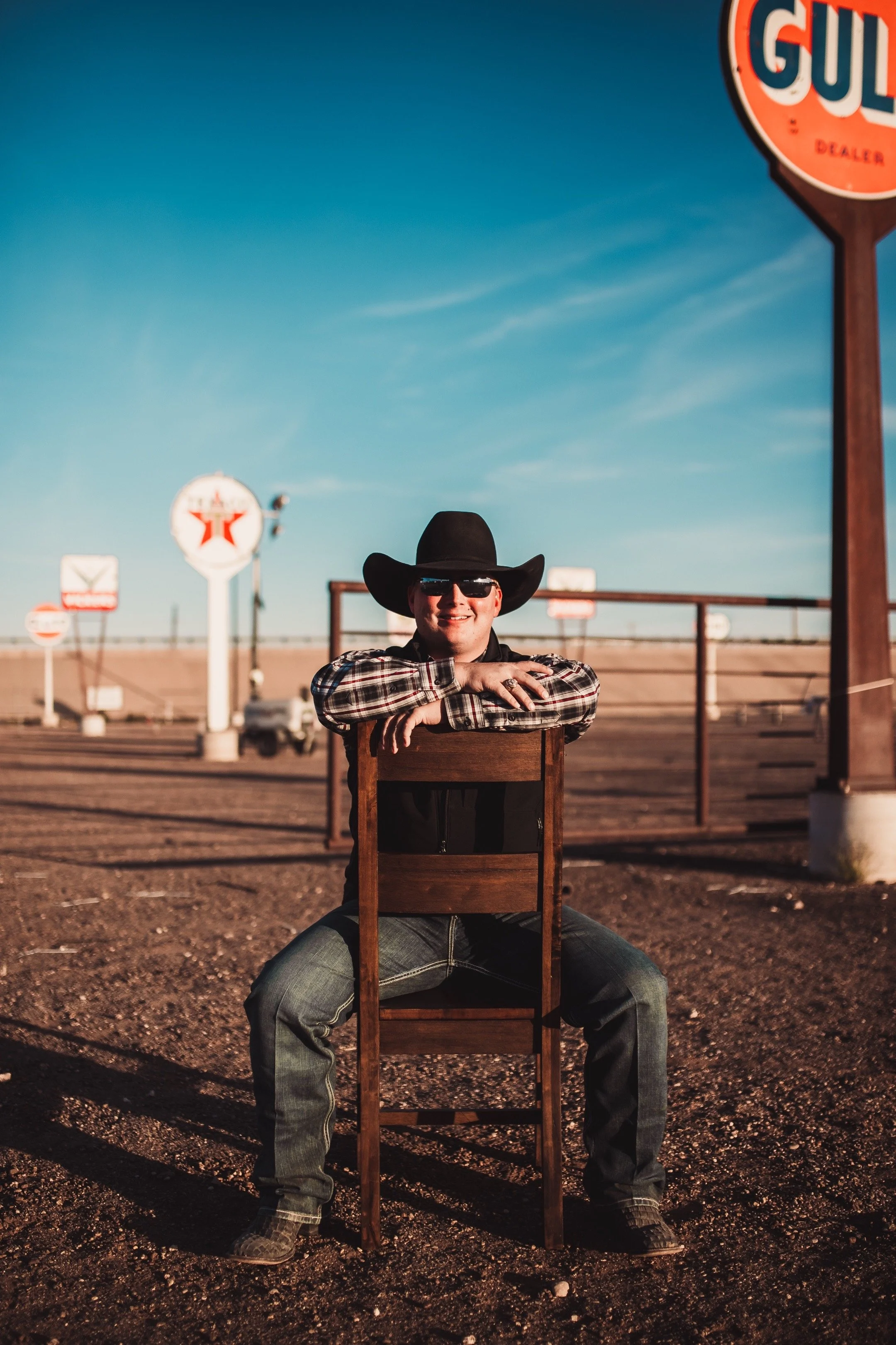 Man wearing cowboy hat and sunglasses sitting on a wooden chair outdoors, with a clear blue sky and Gulf sign in the background.
