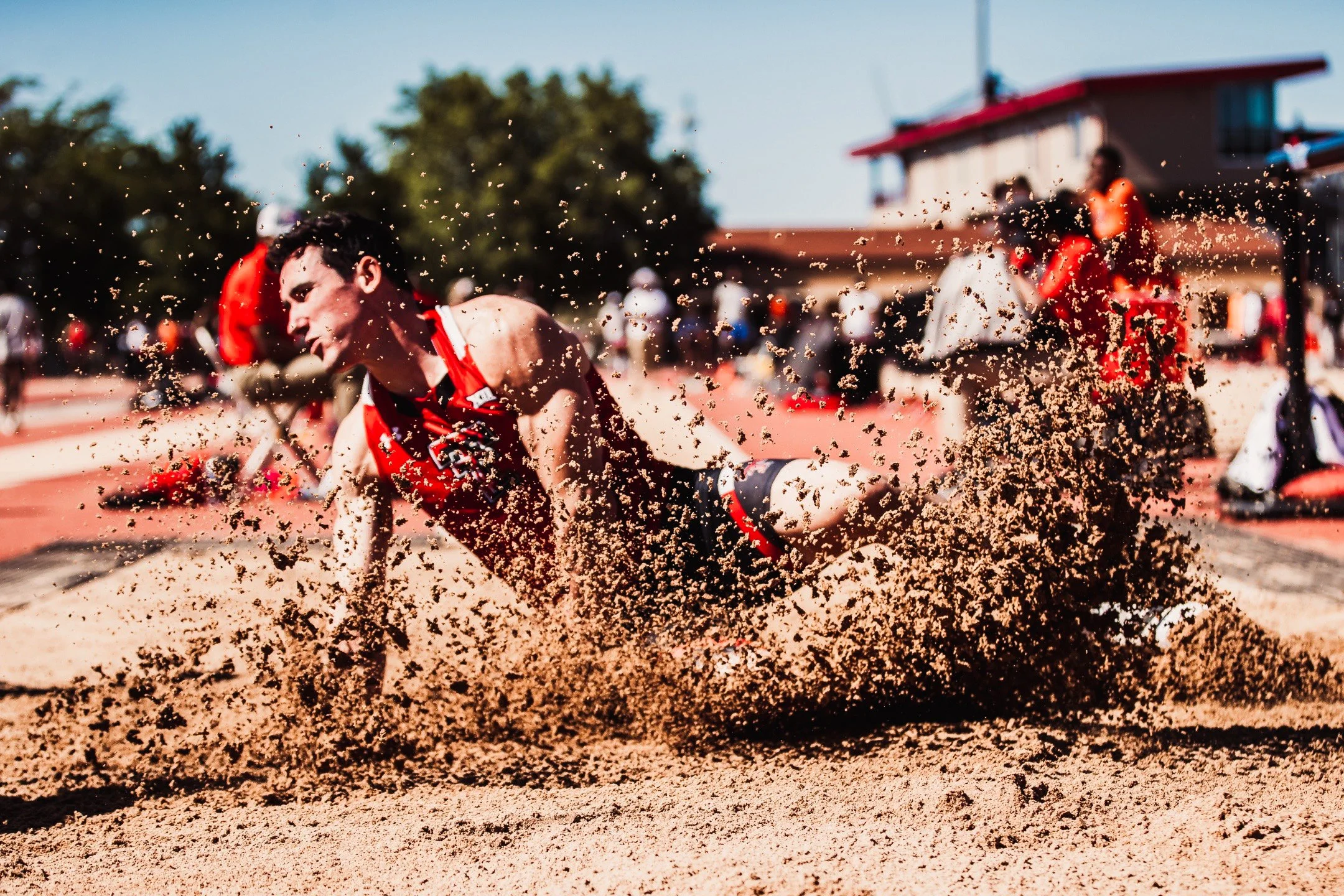 A male athlete in a red uniform falling in a sandpit during a long jump event, with sand flying around him at an outdoor track and field stadium.