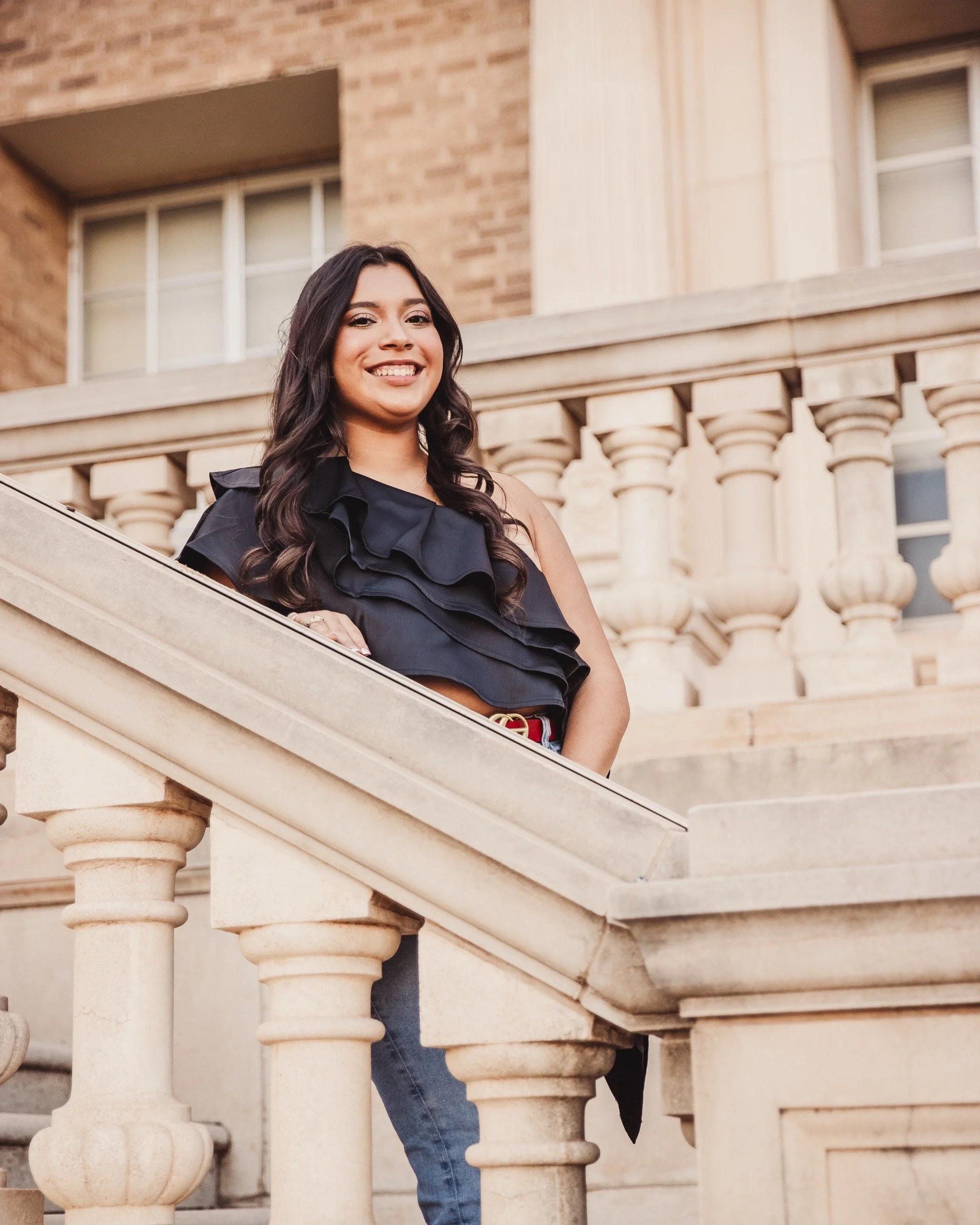 Young woman with long dark hair standing on an outdoor stone staircase, smiling, wearing a black ruffled top and jeans, in front of a building with brick and stone architectural details.